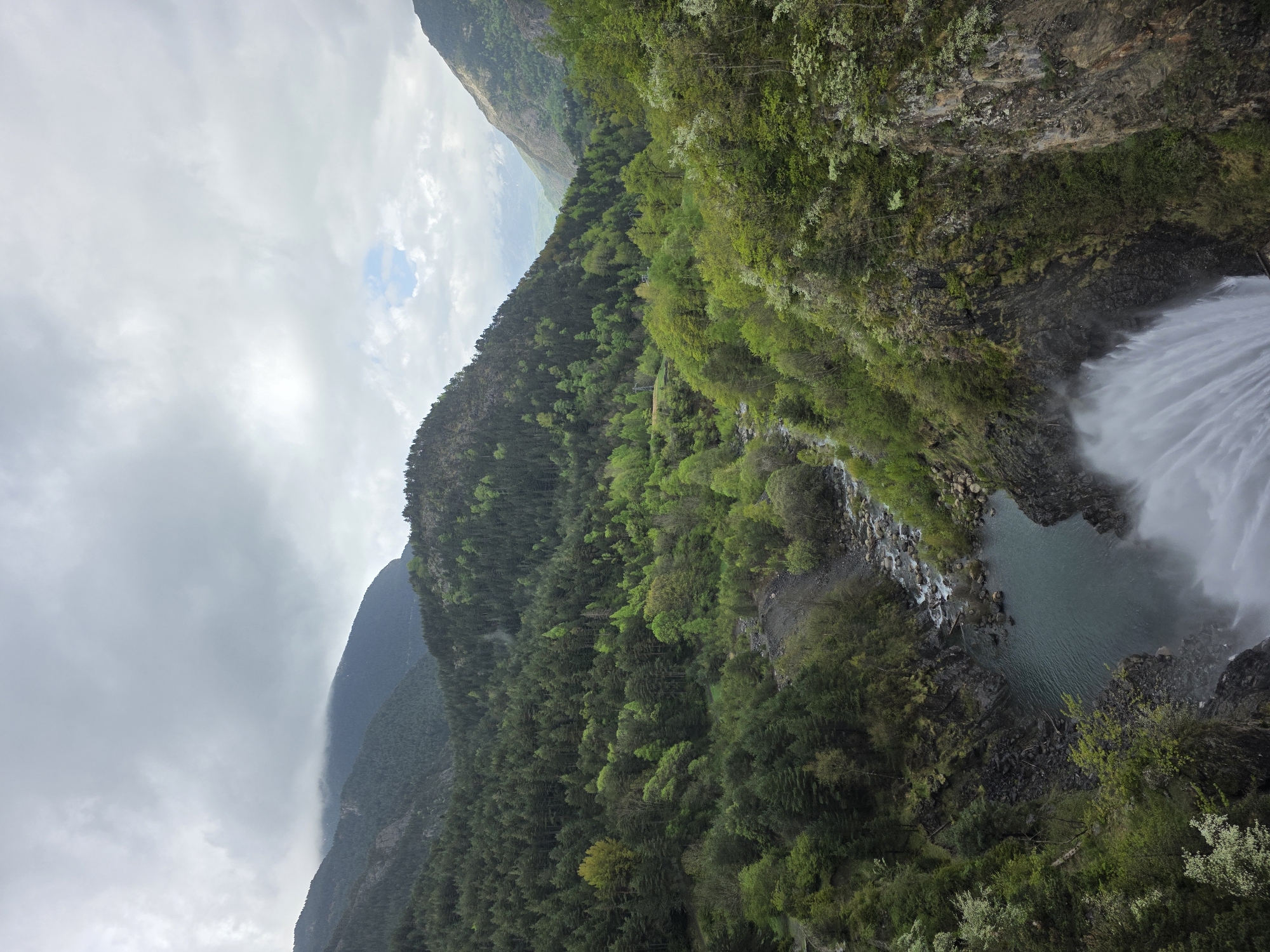 The image displays a stunning natural landscape, predominantly a mountainous and forested area featuring a powerful waterfall. The perspective is from a high vantage point, looking down into a valley.
On the right side of the frame, a large waterfall cascades vigorously down a dark, rocky cliff face. The water appears white and frothy, fanning out as it plunges into a deep, teal-colored pool at its base. Mist rises from the impact of the water, creating a soft atmospheric effect. The pool is surrounded by dark, rugged rocks and smaller rapids and streams that flow into it.
The left and central portions of the image are dominated by steep mountain slopes covered in dense, vibrant green forests. The trees exhibit various shades of green, suggesting lush growth, possibly in spring or early summer. The mountains extend into the background, their peaks partially obscured by a thick layer of grey, overcast clouds. While most of the sky is cloudy, a small patch of brighter blue sky is visible in the upper-left quadrant, hinting at a break in the weather.
No people, animals, or man-made structures are visible, emphasizing the untamed beauty of the natural environment. The scene conveys a sense of grandeur and tranquility, with the raw power of the waterfall contrasting with the stillness of the surrounding forest. The weather appears predominantly cloudy, indicating daylight hours under an overcast sky. This natural landscape is located in Benasque, Spain.