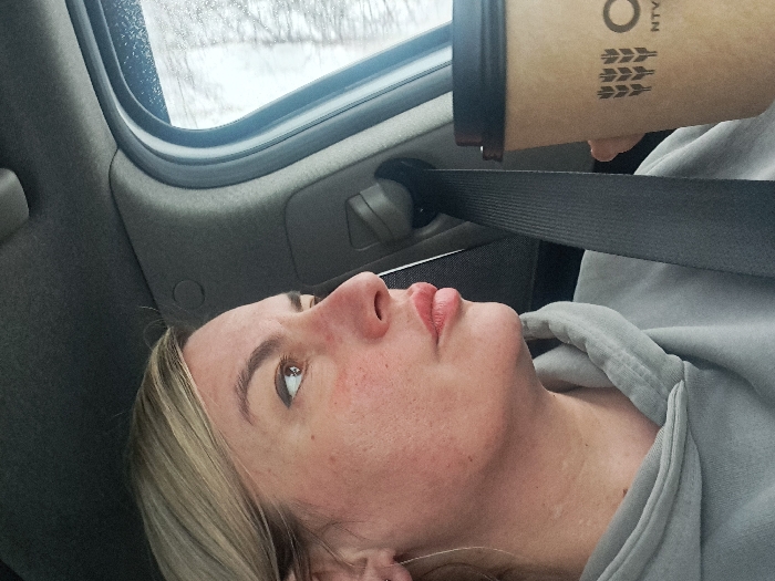 A woman in a gray hooded sweatshirt, wearing a seatbelt, is holding a brown paper coffee cup in a car. The cup is partially visible, displaying a wheat stalk logo. The woman is looking up with a slight smile, her blonde hair partially visible. A car window and interior elements form the background.