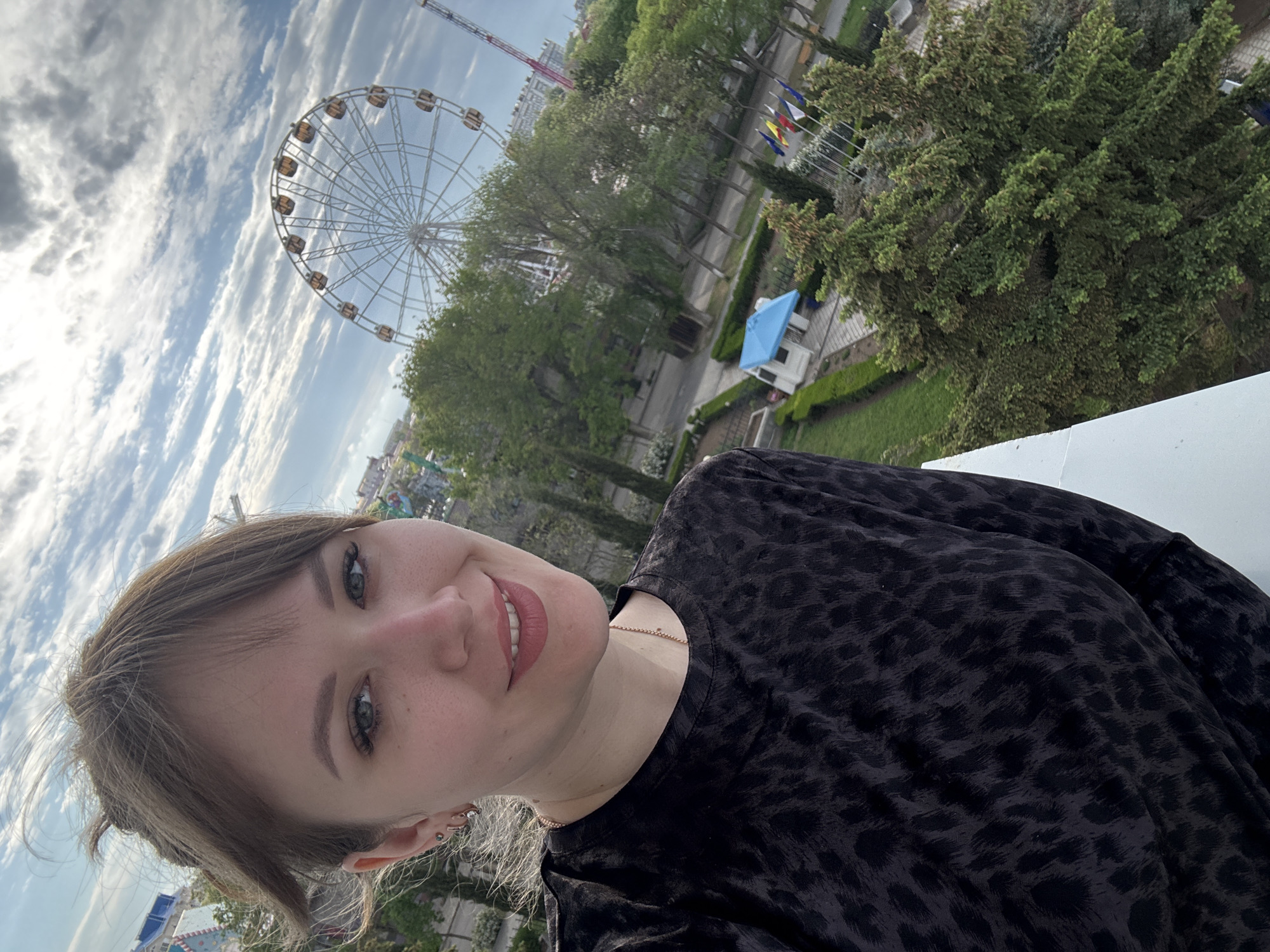 A medium shot depicts a woman in a black patterned shirt, smiling at the camera. Behind her, a Ferris wheel is visible against a cloudy sky. The scene appears to be an amusement park, possibly in Yevpatoriya, Ukraine, based on the Ferris wheel, flags, landscaping, and architecture. A park-like area is seen with trees and bushes, adding to the impression of an outdoor recreation setting.
