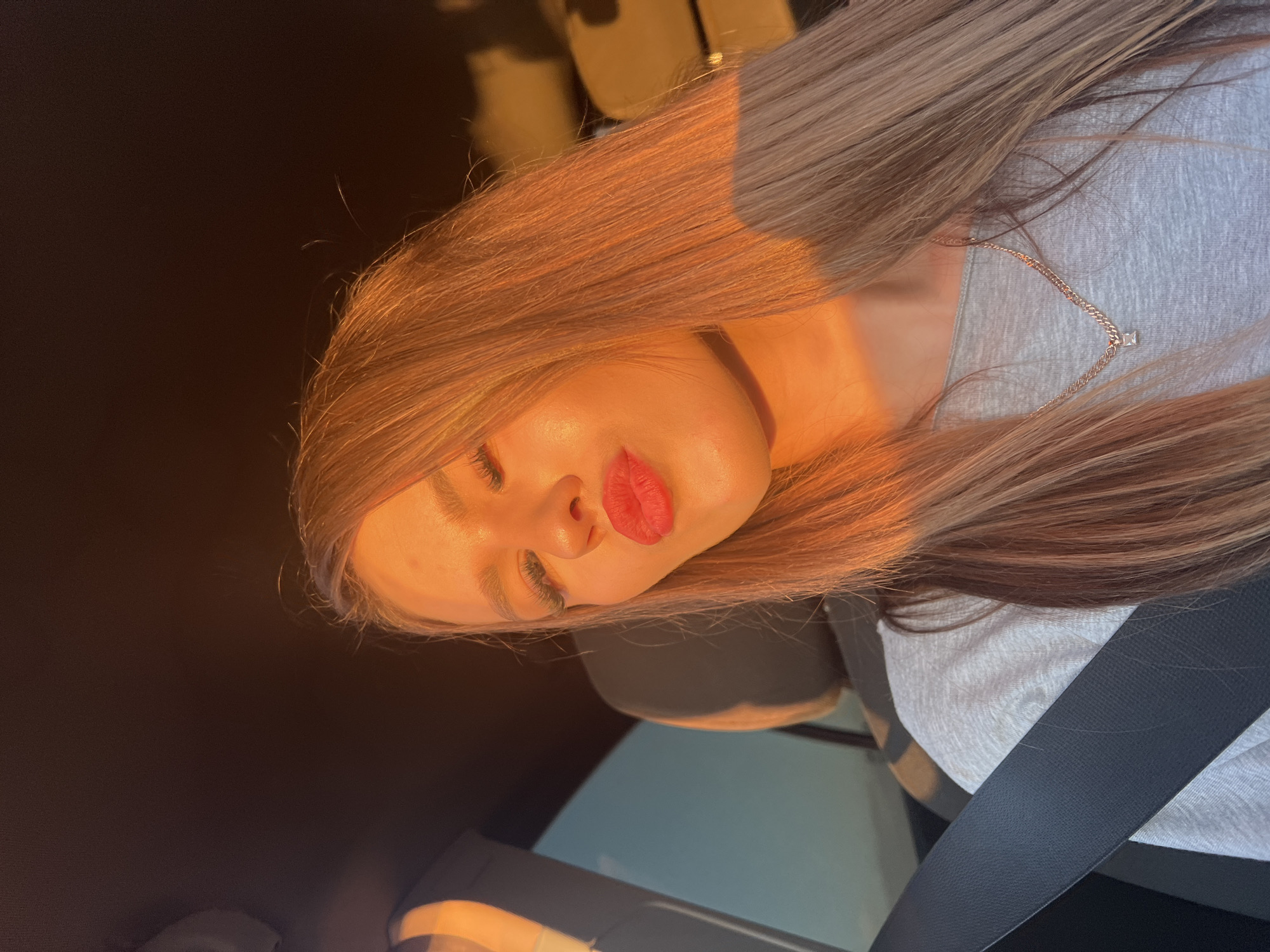 A young woman with blonde hair, wearing a grey t-shirt and a necklace, is pictured inside a car. She is leaning towards the camera, puckering her lips. The setting sun casts a warm, golden light on her face and hair, creating a dramatic effect. A seatbelt is visible across her chest, indicating she is the driver or a passenger. The background shows the interior of the car, with a headrest and the car's ceiling visible. The image does not provide any specific details or landmarks that would allow for an identification of the location within Chaikovsky, Russia.