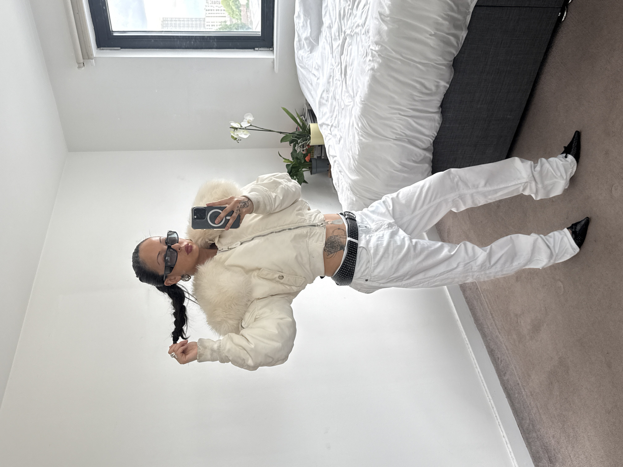 A woman is taking a photo of herself indoors. She has dark hair in a ponytail, sunglasses, a white jacket with a fur collar, white pants, and black shoes. A black studded belt is visible. A tattoo is visible on her torso. She is holding a phone with a circular attachment. The background includes a white wall, a window with a cityscape visible, a bed with white bedding, and a gray headboard. There is also a plant with white flowers on a bedside table. The location is a modern apartment in London, United Kingdom.