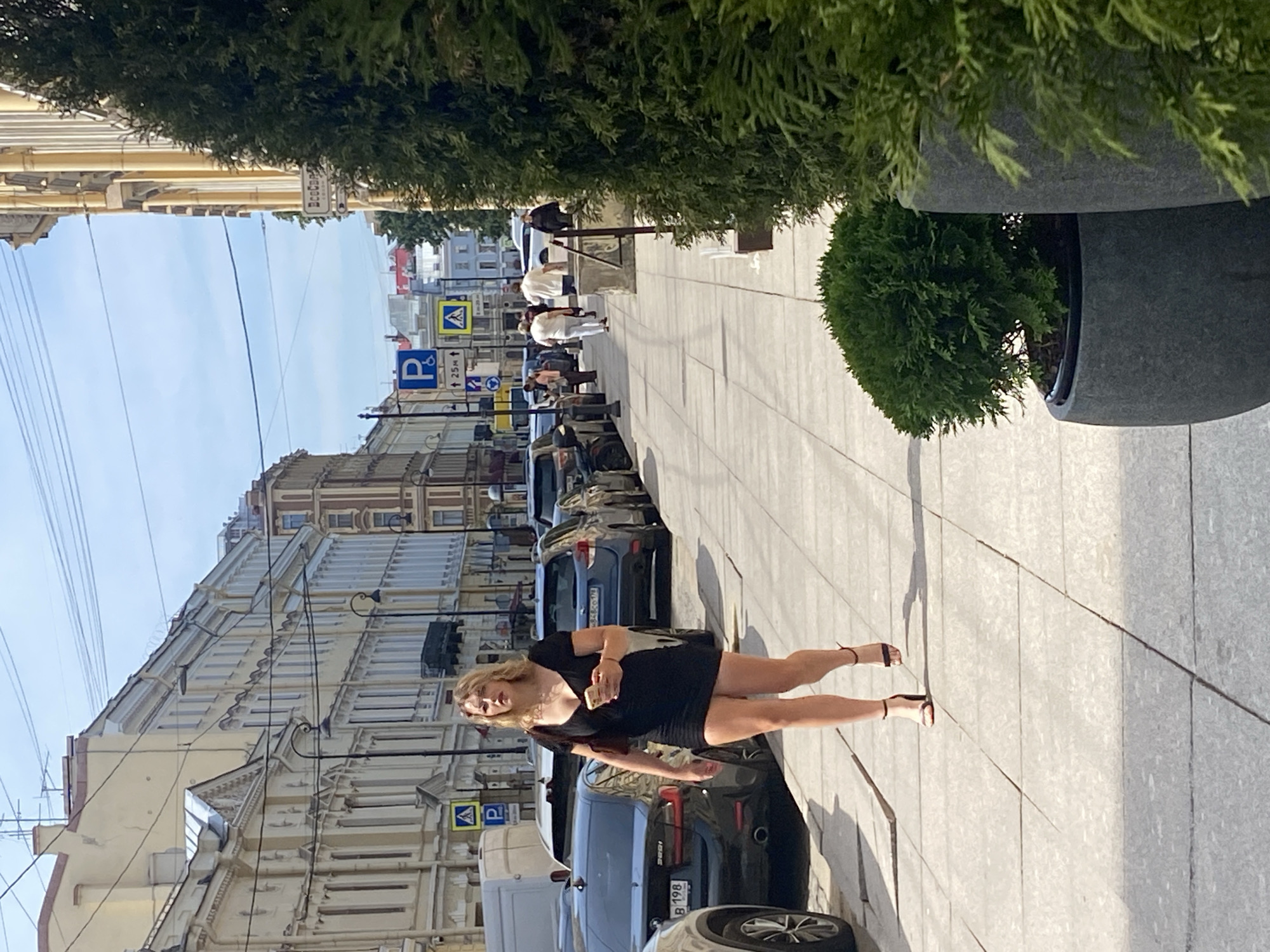 A young woman with blonde hair walks down a sidewalk in Saint Petersburg, Russia. She is wearing a black, short-sleeved, low-cut dress and high-heeled sandals. She is holding a smartphone in her right hand and a dark-colored purse is slung over her left shoulder. The sidewalk is paved with light-colored rectangular tiles. On the left side of the sidewalk, several cars are parked along the street, and buildings with light yellow facades line the street. On the right side of the sidewalk, a large evergreen bush in a grey planter is prominently visible, and further down the sidewalk, other people are walking. The sky is blue with some clouds, and overhead, power lines are visible. A sign with a blue "P" symbol for parking is also visible on the sidewalk. The buildings in the background have architectural details typical of Saint Petersburg.