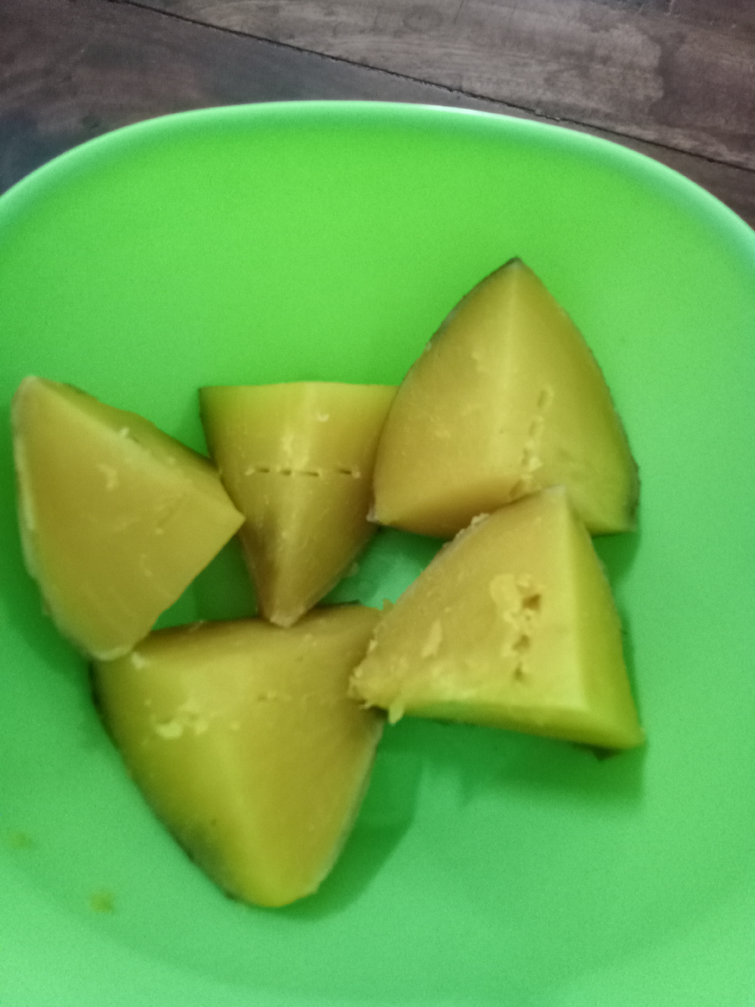 Five triangular pieces of cooked sweet potato are arranged in a bright green bowl, which sits on a wooden surface. The sweet potato pieces are a pale yellow-orange color and appear soft, with some visible pores and slight indentations on their surfaces. The lighting in the image is overhead, casting subtle shadows. The background is out of focus, showing the texture of wood grain. The image was taken at the home of a resident in Simo, Boyolali, Indonesia.