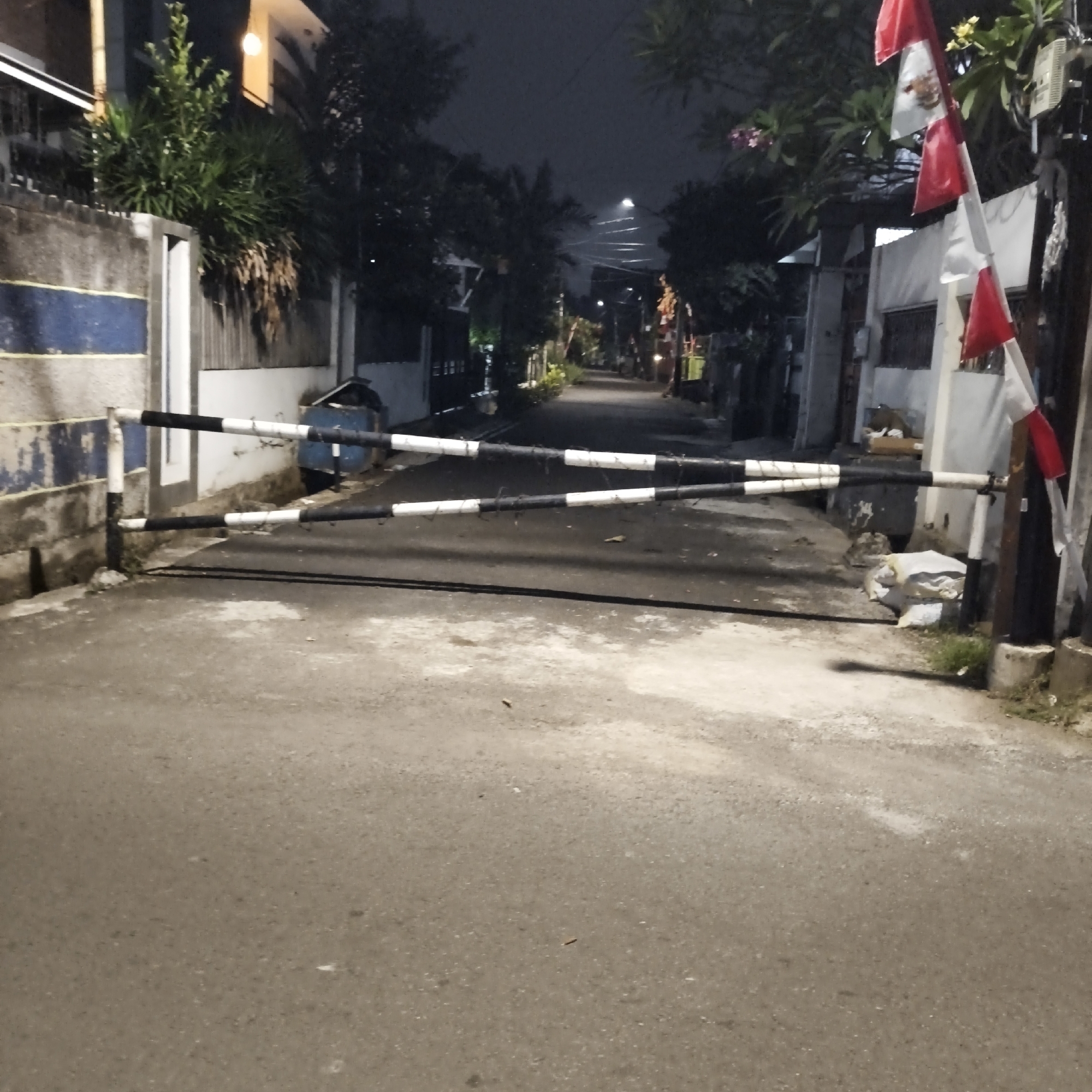 This is a nighttime, medium-shot photograph of a street in South Jakarta, Indonesia. The street is blocked by a horizontal, black and white striped barrier. On the left side of the street is a concrete wall with a blue and white striped pattern. On the right side of the street, a flag pole displays a red and white Indonesian flag. The street is lined with trees and buildings, and a street light illuminates the scene in the distance. There are no people visible in the image. The overall impression is one of a quiet, residential street at night.