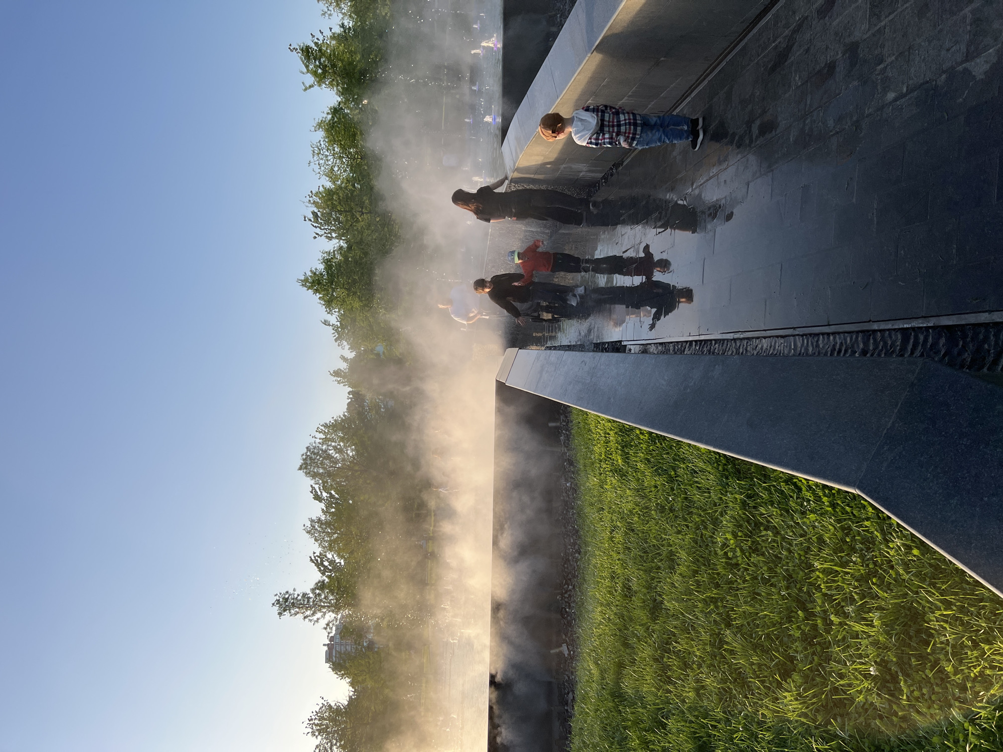 The image depicts a group of people interacting with a water feature in a park. Four figures are visible, appearing to be children and an adult, positioned along the edge of a dark, reflective surface, likely wet stone or tile. Water mist or spray rises in the air around them. The scene is bathed in warm, bright light, suggesting either early morning or late afternoon. The environment includes green grass and trees, with a clear blue sky in the background. The location is identified as the water park in Krasnodar, Russia.