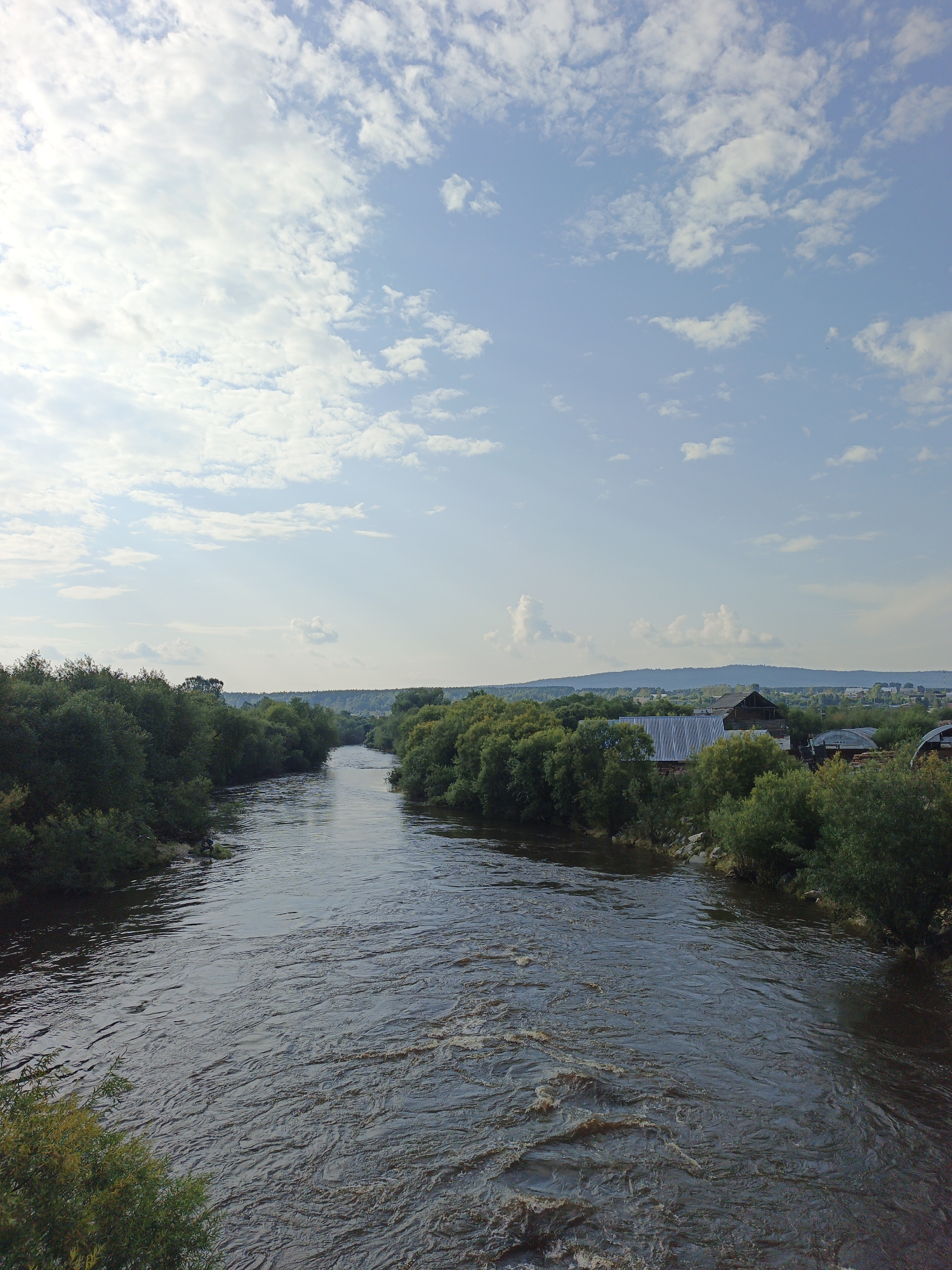 The image is an outdoor, high-angle shot of a river flowing through a rural landscape with a village on the right bank. The river, which appears to be the Ukhta, flows from the background towards the foreground, with visible ripples and some froth suggesting a current. On the right bank, a cluster of houses with corrugated metal roofs and wooden structures are partially visible amidst dense green foliage. In the distance, a range of hills forms the horizon, and the sky is partly cloudy with scattered white cumulus clouds against a pale blue backdrop. The left bank of the river is densely covered with green trees and vegetation. There are no people clearly visible in the image. The overall impression is of a natural, peaceful rural setting.