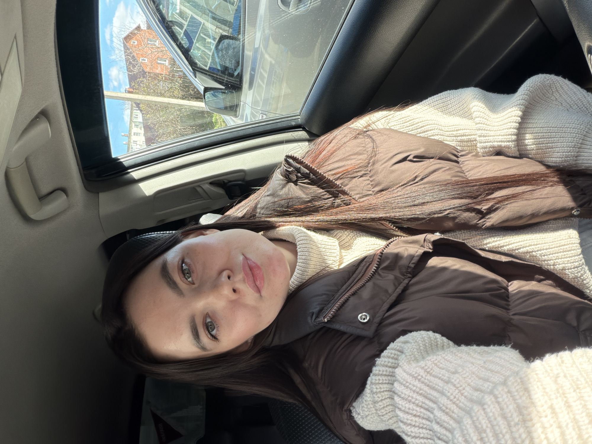 A young woman is featured in a close-up, horizontal selfie taken from inside a car. She has long, straight dark brown hair, light-colored eyes (appearing greenish-grey), and full, lightly tinted lips. She wears a cream or off-white ribbed turtleneck sweater beneath a dark brown puffer jacket.
She is seated in a car, likely in a passenger seat, with bright sunlight streaming in through the window on her right side, casting highlights on her hair and jacket. The interior of the car, including parts of the dashboard, door panel, and headliner, is visible around her.
Outside the car window, an urban scene is illuminated by a clear, sunny sky with some scattered clouds. A prominent reddish-brown multi-story building is visible, partially obscured by bare tree branches, suggesting a late autumn or early spring season. Another lighter-colored residential building can be seen in the distance, and reflections in the window indicate the presence of other vehicles.
The time of day is bright daylight, indicating midday or afternoon. The woman maintains a neutral, calm expression while looking directly at the camera. There is no discernible text visible within the image. The provided location context for this scene is Chaikovsky, Russia.