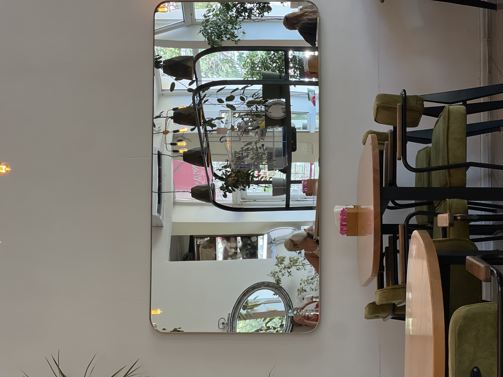 The image captures a wide rectangular mirror mounted on a white wall, reflecting the interior of a cafe. In the foreground, two round wooden tables with dark metal legs are partially visible, accompanied by green upholstered chairs with wooden armrests. A wooden box filled with pink sugar packets sits on one of the tables.
The mirror's reflection reveals a bustling cafe environment. Several people are visible, including a woman with blonde hair in a ponytail sitting at a table, and another woman taking a photo with her phone. Behind them, shelves are stocked with various items. Plants are strategically placed throughout the cafe, adding a touch of greenery. A birdcage hangs from a shelf, suggesting a pet-friendly atmosphere. Light fixtures with Edison bulbs hang from the ceiling, casting a warm glow. Large windows in the background allow natural light to stream in, and outside, trees are visible.
The cafe appears to be located in the city of Odesa, Ukraine, as suggested by the text visible in the reflection.
