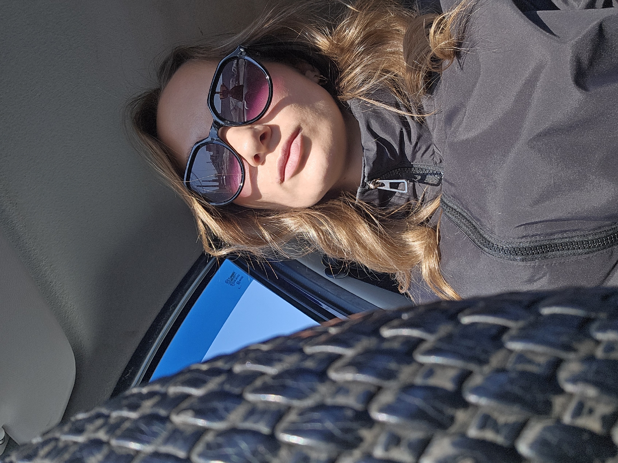 A person is lying down, looking up at the camera, wearing sunglasses and a black jacket. There is a car window above and to the left of the person, showing a bright blue sky. Below the window, there is a close-up view of a patterned object, possibly a tire or a textured surface. The photo is taken from an unusual angle, suggesting it may have been taken inside a vehicle. The location in the city of Chaikovsky, Russia is not discernible from the visual information provided.