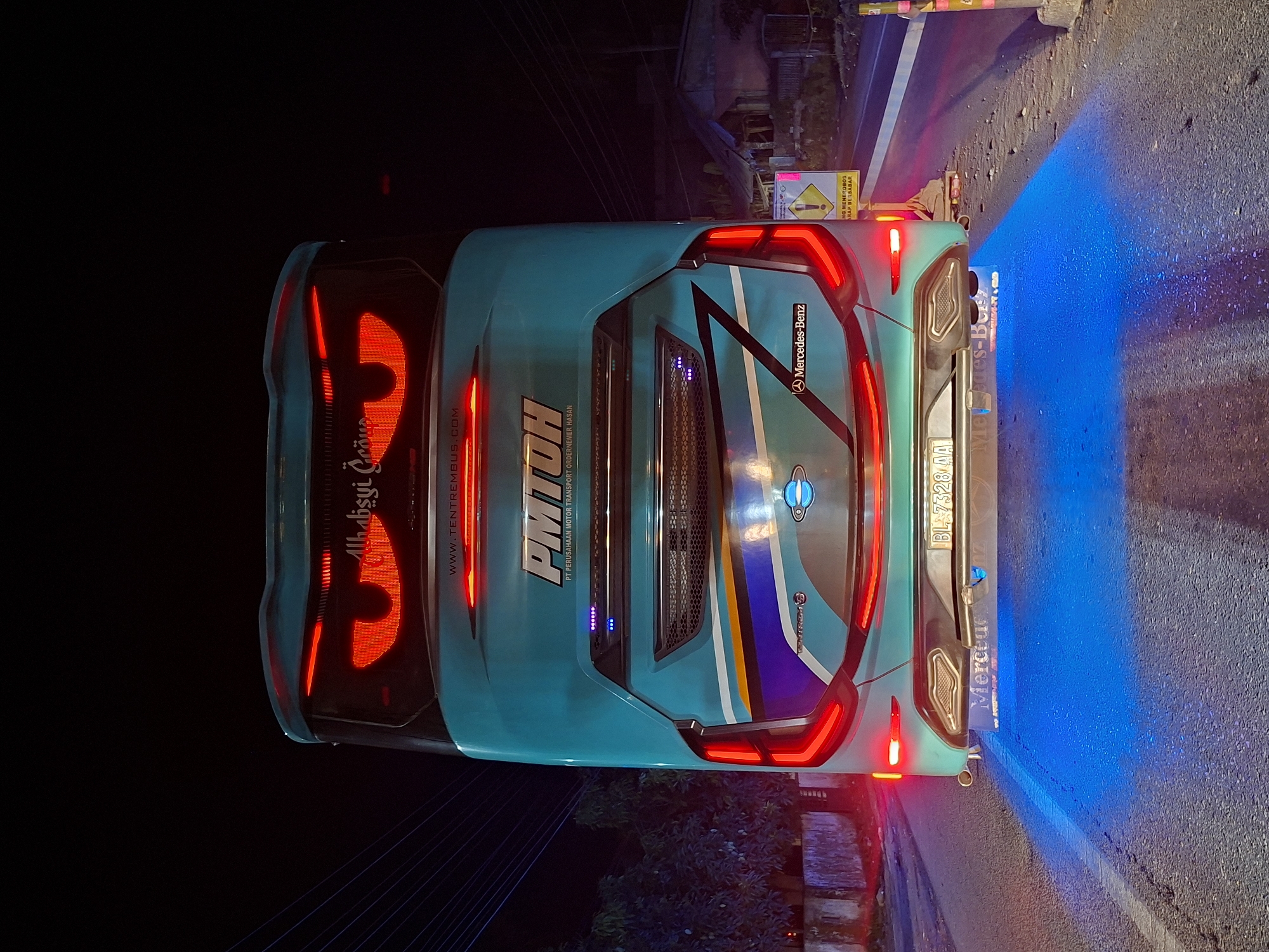 The image depicts the rear of a light blue PMTOH bus at night on a road. The bus, identified by a Mercedes-Benz logo, displays the license plate BL 7328 AA. The bus's tail lights are illuminated, and blue light emanates from beneath it, reflecting on the road. The text "Ahabsyi Group" and "WWW.TENTREMBUS.COM" are visible on the left side of the bus. A road sign written in Indonesian is visible on the right. The bus's logo also appears to include a depiction of a planet. The name of the city Lhokseumawe, Indonesia is not visible in the provided image.