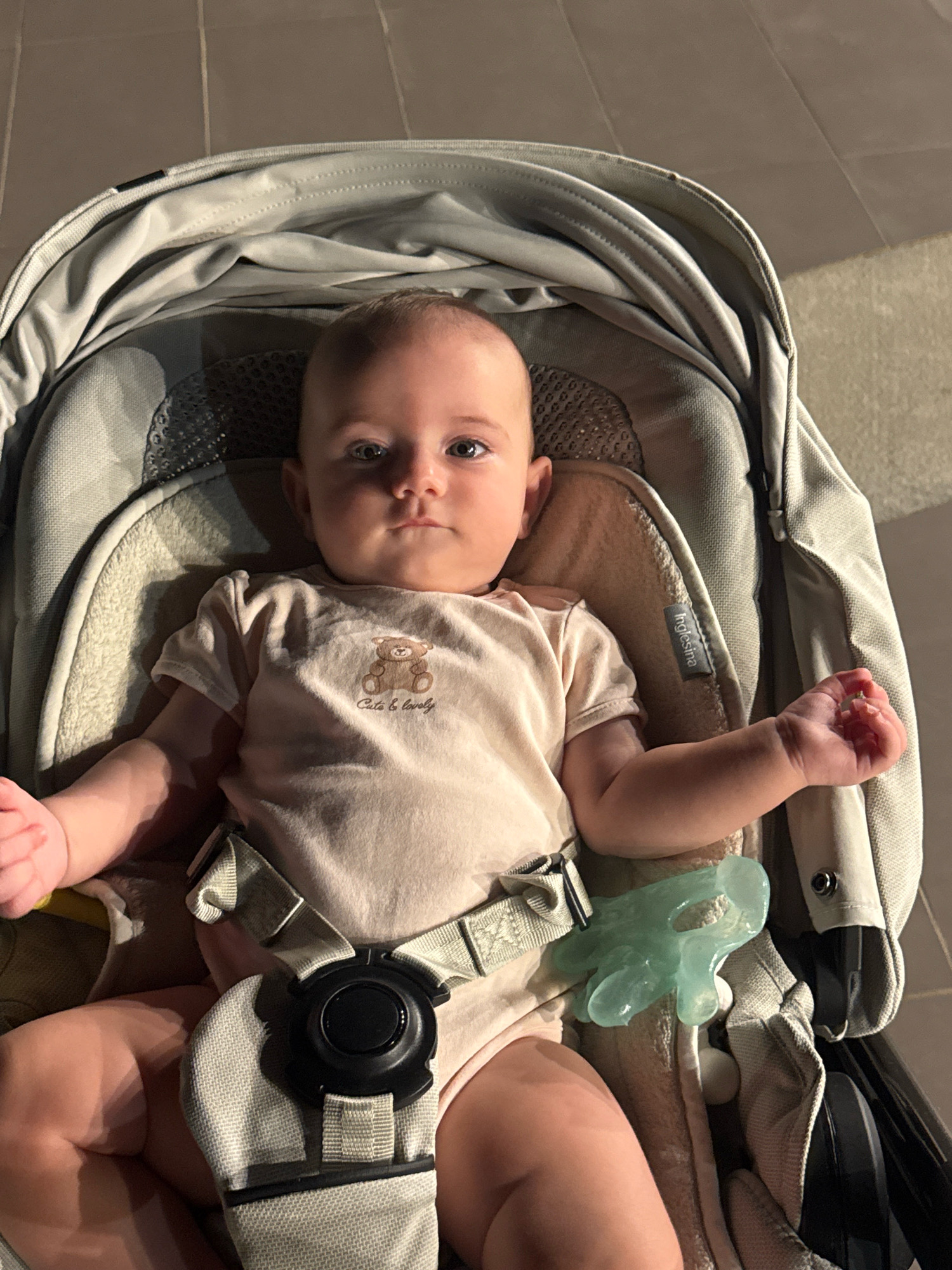 A baby is lying in a stroller, looking towards the camera. The baby is wearing a light pink onesie with a teddy bear graphic and the words "Cute & Lovely" printed below it. The stroller is gray and has a five-point harness securing the baby. A light green pacifier is attached to the side of the stroller. The background appears to be an outdoor setting with tiled flooring. The lighting suggests it is evening or night, with artificial lights casting shadows. The location is the city of Isola delle Femmine, Italy.