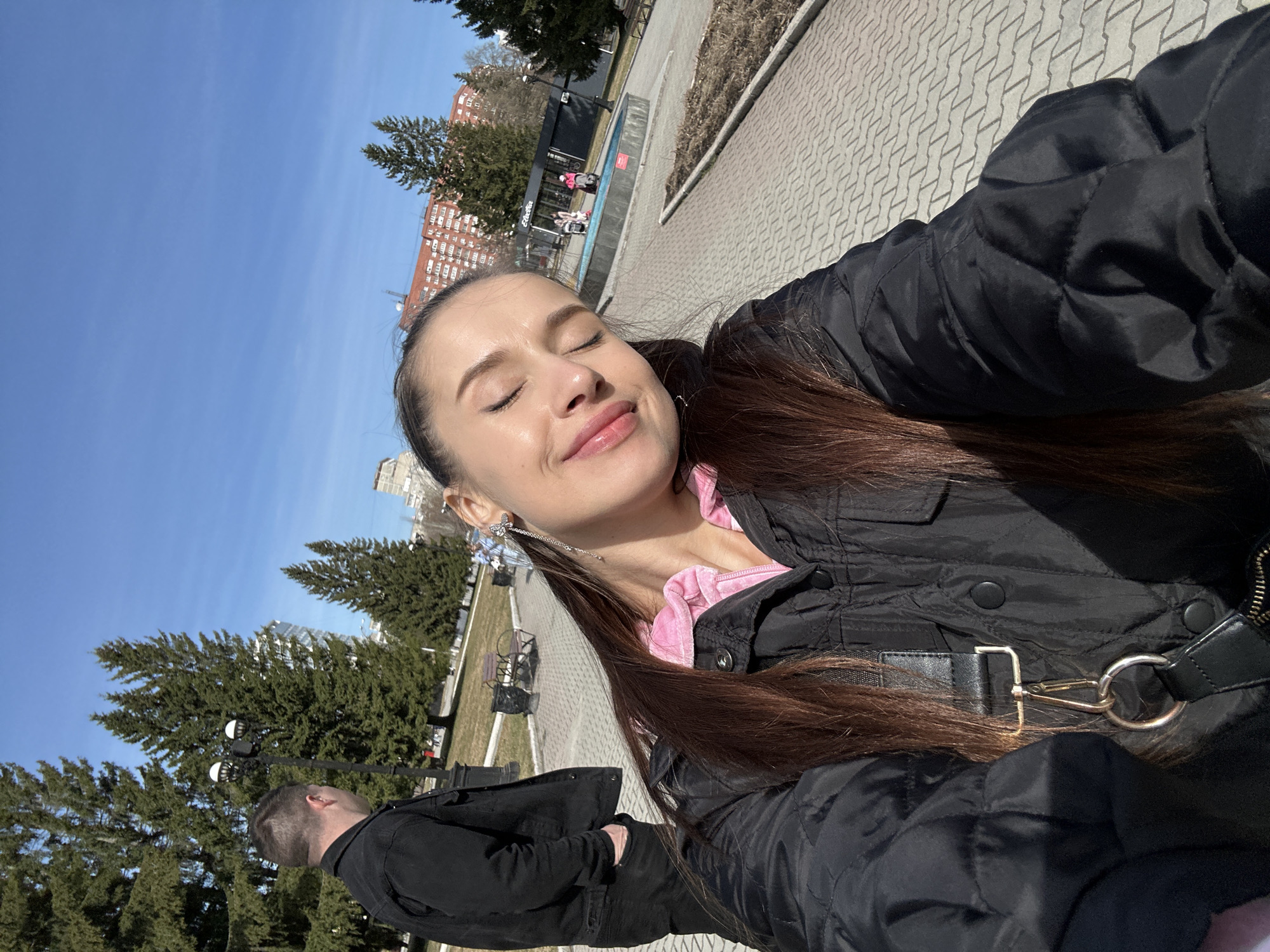 A selfie taken in Yekaterinburg, Russia shows a young woman outdoors on a sunny day, with her eyes closed, seemingly enjoying the sun. She is wearing a black jacket over a pink top and dangling earrings. A man in a black jacket walks in the background. The ground is paved with bricks. There are trees, benches, and buildings in the background, typical of a city park or square. A structure that might be a bus stop or small shop can be seen in the far background.