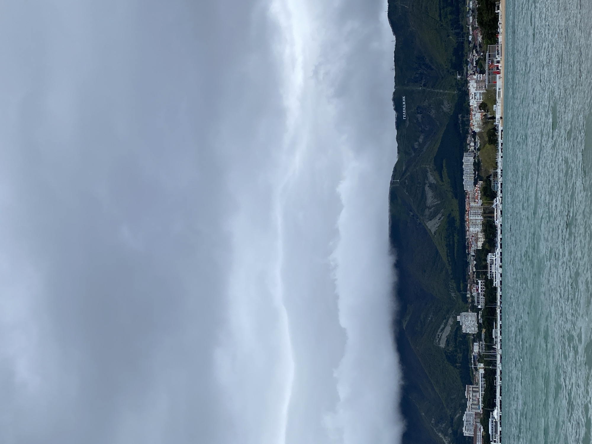 The image depicts a coastal scene in Gelendzhik, Russia. On the right, calm turquoise water meets the shore. Adjacent to the water is a row of buildings and structures extending along the coastline. Behind the buildings, a green mountain rises, bearing the inscription "ГЕЛЕНДЖИК" in large white letters on its slope. The left side of the image is dominated by a gray, overcast sky with thick cloud formations. The foreground elements, such as buildings and the shoreline, are clearly visible, contrasting with the more obscured mountain and sky.