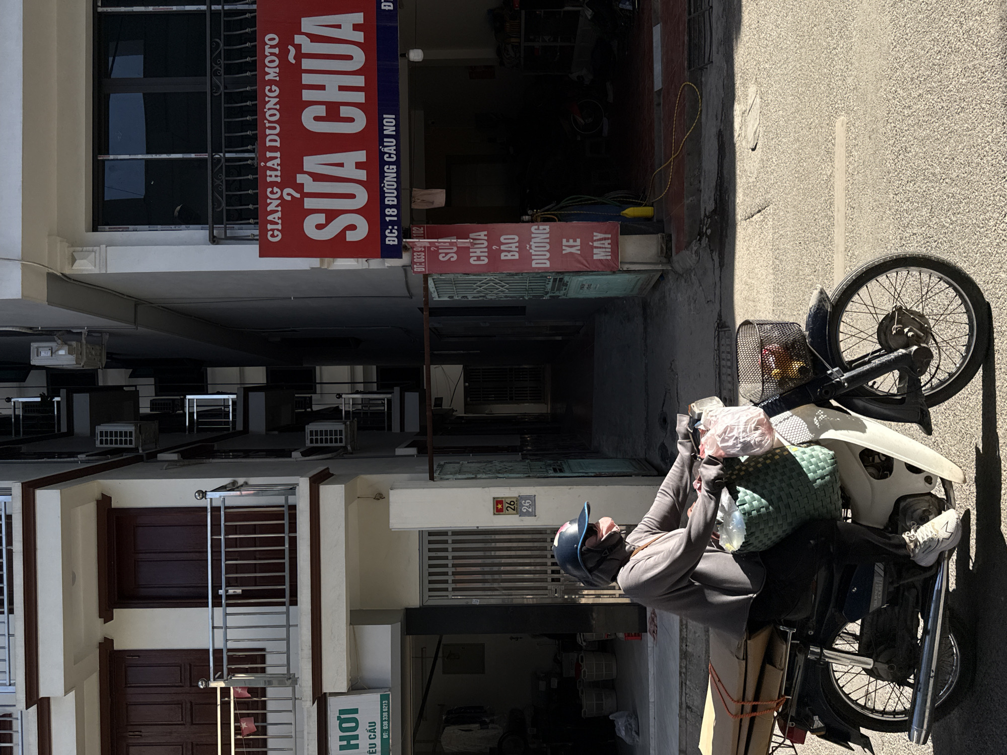 The image shows a street scene in Hà Nội, Vietnam, with a person on a scooter parked near a motorcycle repair shop. The repair shop's sign reads "GIANG HẢI DƯƠNG MOTO SỬA CHỮA" which translates to "Giang Hai Duong Motorcycle Repair". The address is "ĐC: 18 ĐƯỜNG CẦU NOI", meaning 18 Cau Noi Street. The person on the scooter is wearing a helmet and casual clothing. The scooter has items loaded in the front basket and on the seat. Buildings line the street with number 26 displayed on one facade and "HƠI ÊU CẦU" sign on another facade. The street surface is asphalt. The time of day appears to be daytime with harsh sunlight.