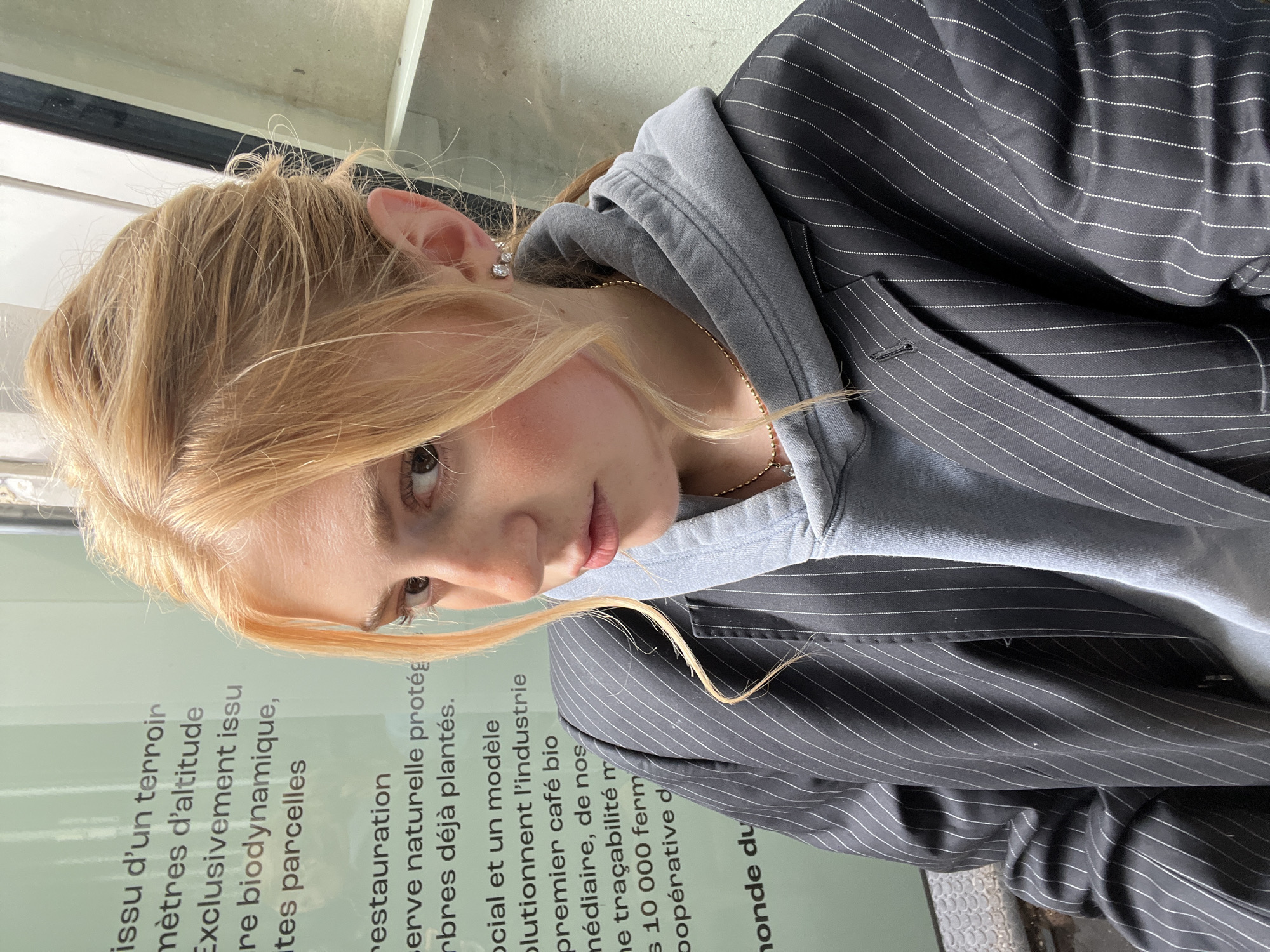 A young woman with blonde hair poses close to a sign in Paris, France. She wears a grey hoodie under a black pinstriped blazer and a delicate gold necklace. The sign next to her contains French text relating to organic farming. The shot is taken at an unusual angle.