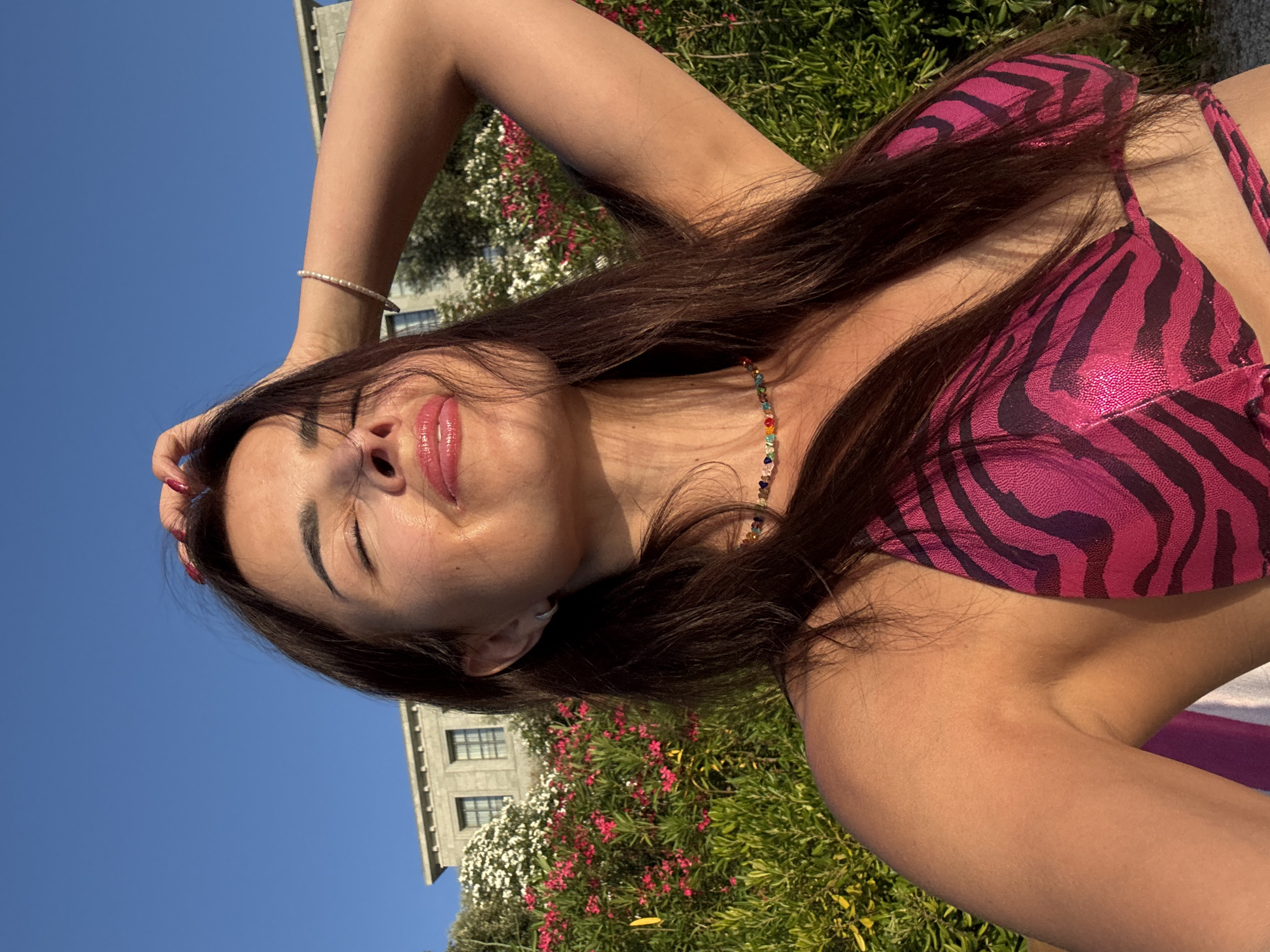 This is a close-up, low-angle selfie of a young woman lying down outdoors, likely in a grassy area with blooming flowers, possibly a garden or park. She is wearing a bright pink bikini with a black zebra-like print. The sun is shining, casting highlights on her face and skin. Her eyes are closed, and her lips are slightly parted, suggesting a relaxed or sensual expression. She has long, dark brown hair that is partially covering her face and shoulder. A colorful beaded necklace is visible around her neck. Her right arm is raised and bent, with her hand behind her head. A delicate pearl bracelet adorns her left wrist. In the background, out of focus, are green bushes with small pink and white flowers, a building with windows, and a clear blue sky. The overall mood is casual and summery, evoking a sense of leisure and enjoyment of the outdoors. No visible text is present.