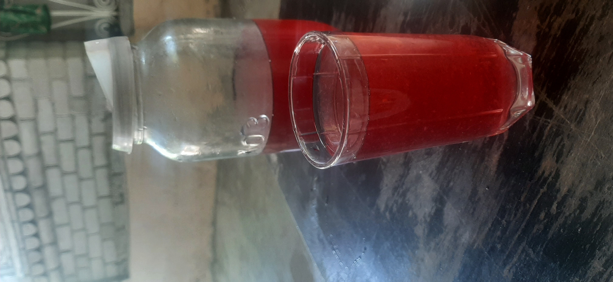 This is a close-up, landscape-oriented photograph of a clear glass jar and a drinking glass filled with a red liquid, likely a beverage. The jar appears to be partially filled with a clear substance, possibly water, and has a white plastic lid. There is some condensation on the outside of the jar, suggesting it is cold. The jar has a circular indentation with the number "30" visible on its surface.
Next to the jar is a clear glass tumbler also containing the same red liquid. The glass has a rim with a textured edge. The subjects are placed on a dark, reflective surface that appears to be a table. The background on the left side of the image shows a light-colored, textured surface that resembles a tiled wall or a brick pattern, with some green and white elements visible at the very top left edge.
The overall lighting suggests it might be daytime, possibly indoors with natural light. There are no people visible in the image. The scene is still and focuses solely on the containers and their contents. No specific event is taking place; it appears to be a simple arrangement of drinkware. No discernible emotions are conveyed, and there is no visible text other than the "30" on the jar. The location context provided is Yessentukskaya, Russia.