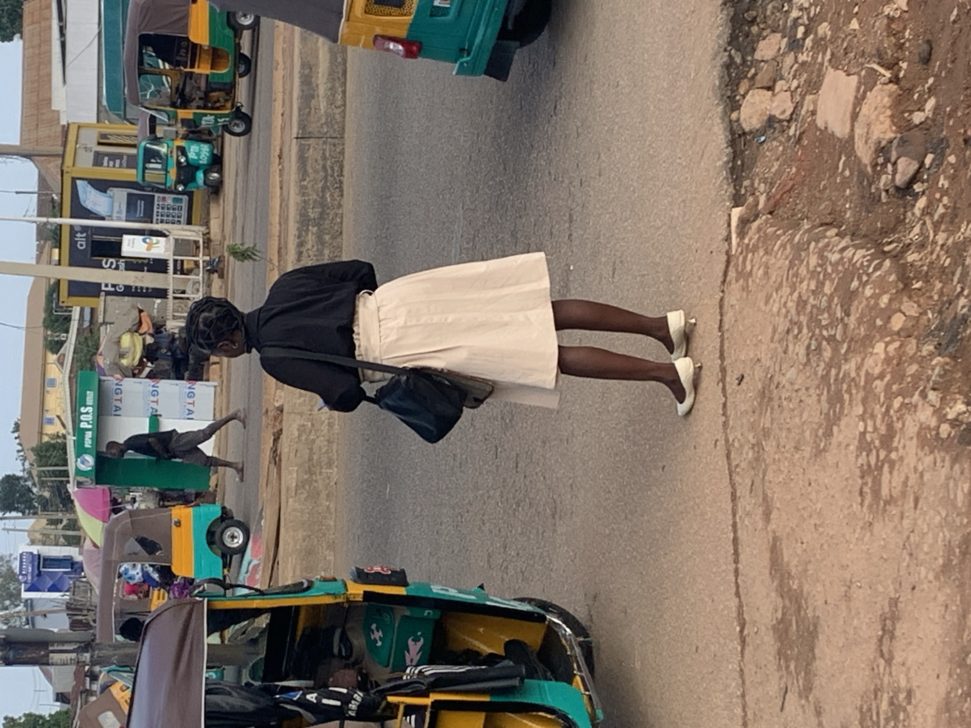 In Gura Top, Nigeria, a Black woman with her hair in braids is standing on a dusty road, facing away from the camera. She is wearing a black long-sleeved shirt and a light-colored, knee-length skirt. Her feet are clad in white ballet flats with a small heel. She carries a dark-colored shoulder bag on her left side.
The road is paved and shows signs of wear, with patches of dirt and debris. In the background, a bright yellow and green auto-rickshaw (also known as a "keke") is parked on the left side of the frame. Further back, another similar auto-rickshaw, primarily green and yellow, is visible with passengers inside.
Across the street, there are structures that appear to be market stalls or small shops. One prominent stall has a green facade with "P.O.S" written in white letters. Next to it, a yellow shipping container-style structure bears the "alt" logo and images of payment cards and a keypad. A man in a dark shirt and shorts is walking towards the P.O.S. stall. The overall environment suggests a busy urban street scene with commercial activity. The sky is overcast, and the lighting indicates it is daytime.