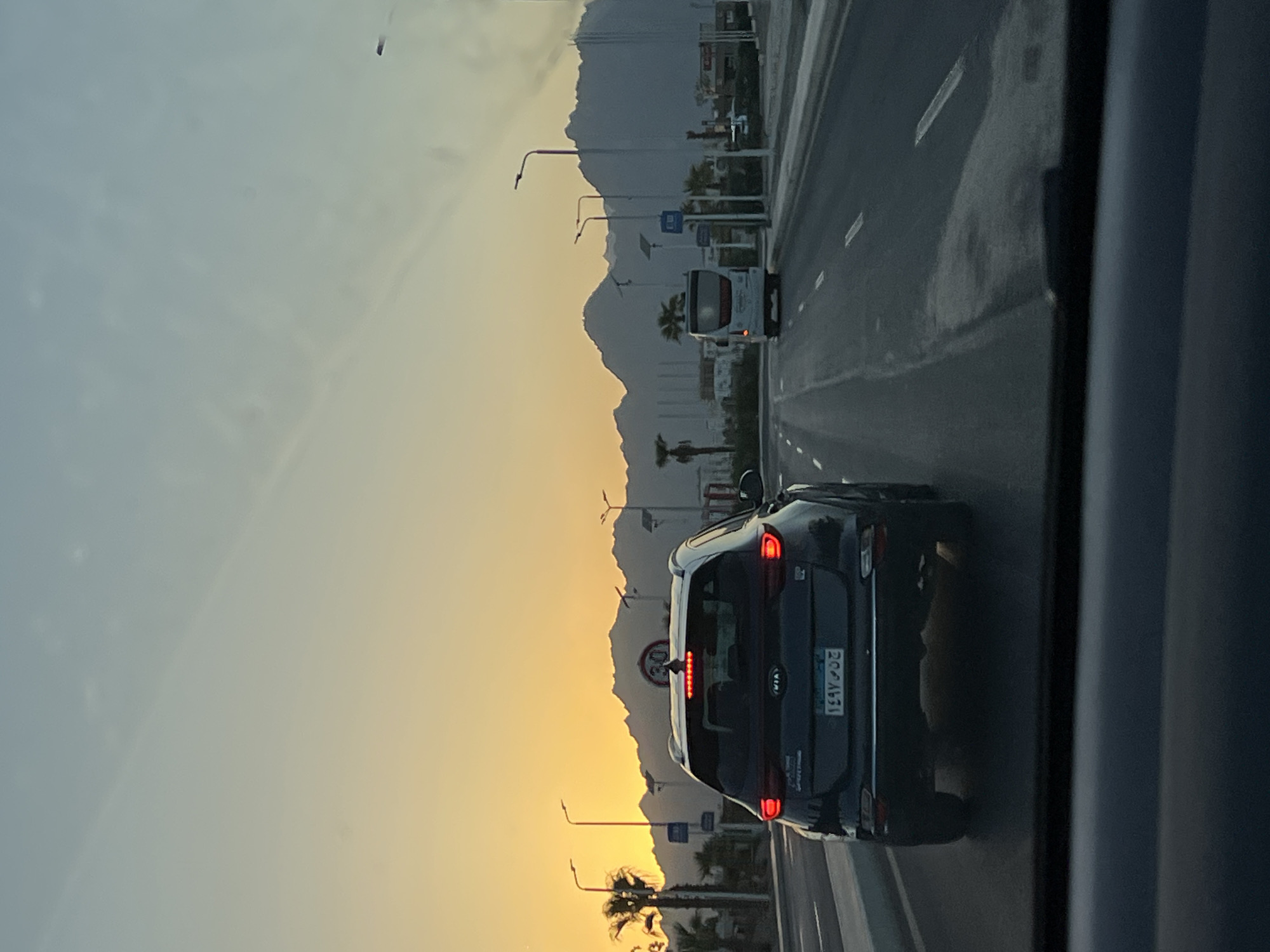 The image captures a view from inside a vehicle driving on a multi-lane road in Sharm El Sheikh, Egypt, during sunset. The sky displays a gradient of soft blues and oranges, with the sun low on the horizon behind a range of mountainous terrain. Palm trees and streetlights line the sides of the road. In the foreground, the rear of a dark-colored SUV is visible, its brake lights illuminated. Further ahead on the road, a white bus is traveling in the same direction. Traffic signs are present along the roadside, including a circular sign with "30" indicating a speed limit. The road is paved and appears to be in good condition, with clear lane markings. Buildings and structures are visible in the distance, suggesting an urban or developing area. The overall impression is a peaceful, scenic drive at dusk.