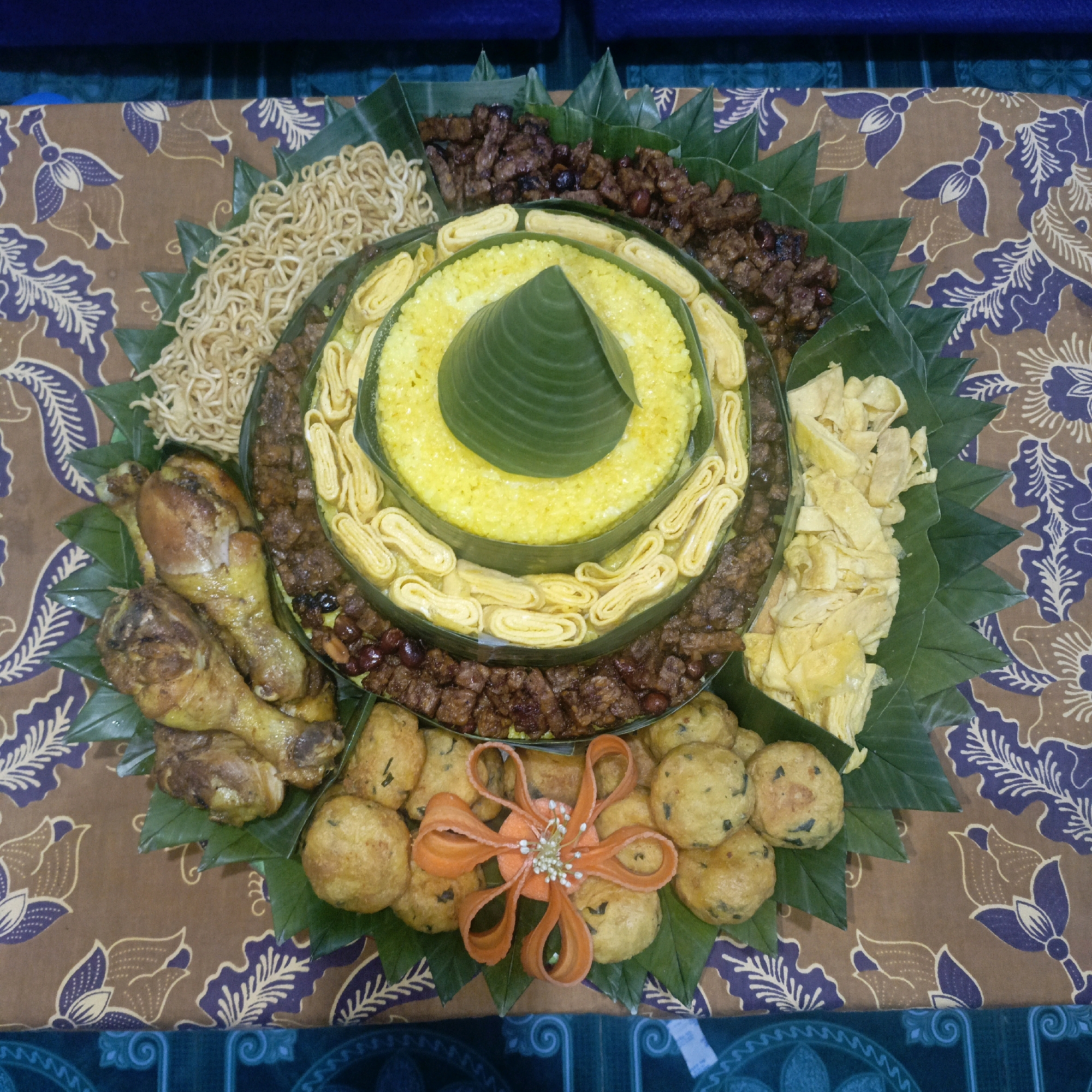 A circular platter of Indonesian food, likely a Nasi Tumpeng, is displayed on a brown and purple batik cloth. The platter is arranged with layers of food on banana leaves. The central element is bright yellow rice, shaped into a cone and topped with a green banana leaf. Surrounding the rice are rings of yellow omelet slices and dark brown pieces of meat. Further out, there are piles of fried noodles, fried chicken drumsticks, and small, round fried fritters. A decorative element made from carved orange carrots and small white seeds is placed in the center of the fritters. The platter is positioned on a table covered with a batik fabric that has a brown base and a repeating pattern of purple floral motifs. In the background, a glimpse of dark blue upholstery is visible. The scene appears to be set indoors, possibly for a celebratory meal. The context of Salatiga, Indonesia, suggests a traditional Indonesian culinary presentation.