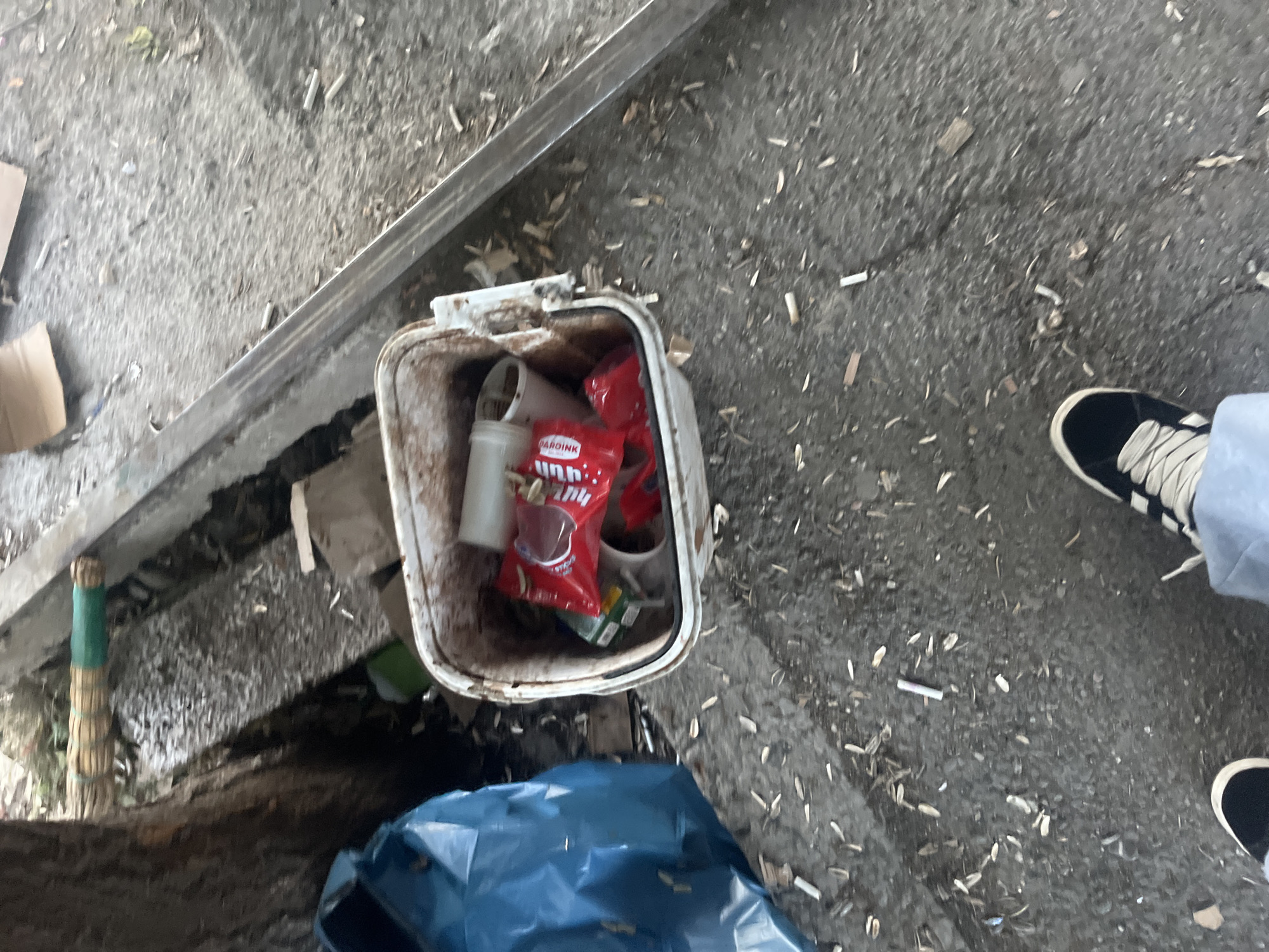 The image depicts a street scene in Yerevan, Armenia, showing a discarded white plastic trash can overflowing with litter, including a package of "Bardink" sunflower seeds. The trash can sits on a concrete surface littered with cigarette butts and small pieces of wood debris. A blue plastic garbage bag partially obscures the base of the trash can. A broom with a green handle lies adjacent to the trash can. A person's legs are visible, wearing black and white sneakers. A metallic rail or curb runs diagonally across the upper left of the frame.