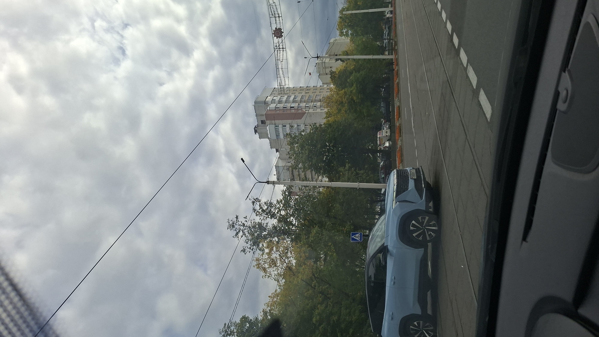 The image shows a light blue SUV driving on a city street in Vitsebsk, Belarus. The street is paved and has traffic lanes marked with white lines. There are tram tracks visible in the road. On the right side of the road, there are trees and a tall apartment building. The sky is overcast with clouds. A pedestrian crossing sign is visible to the left of the road. The photo appears to be taken from inside another vehicle, as the dashboard and windshield are visible in the foreground.
