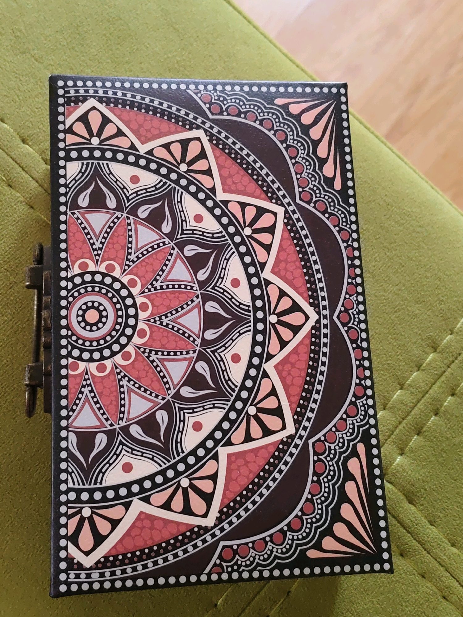 The image shows a closed, decorative box with a mandala design on its lid. The box is dark brown with intricate patterns in shades of pink, light gray, white, and black. The mandala design features concentric circles and petal-like shapes. A small, antique-style metal latch is visible on the left side of the box. The box is resting on a textured green surface, possibly upholstery. The background is blurred but suggests an indoor environment with wooden flooring. The scene is static and shows no people or activity. The location is not identifiable from the visual information provided.