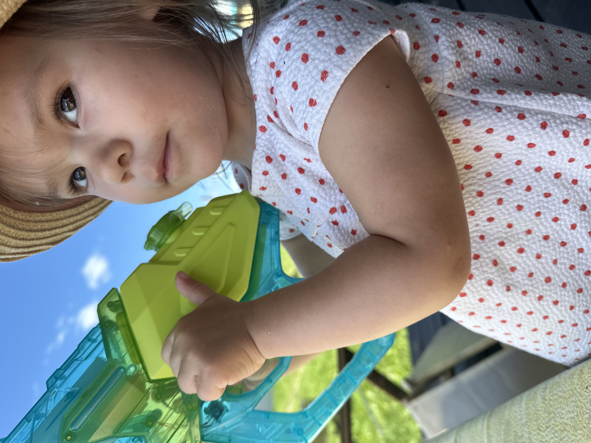 A close-up shot shows a young child wearing a straw hat and a white dress with red polka dots, holding a blue and green water gun. The child is looking upwards, to the right of the frame. The background consists of a bright blue sky with some clouds and green foliage, suggesting an outdoor setting, potentially a backyard in Kharnavy, Russia. The child's hand is gripping the water gun firmly. The image focuses on the child and the toy, with the environment blurred.