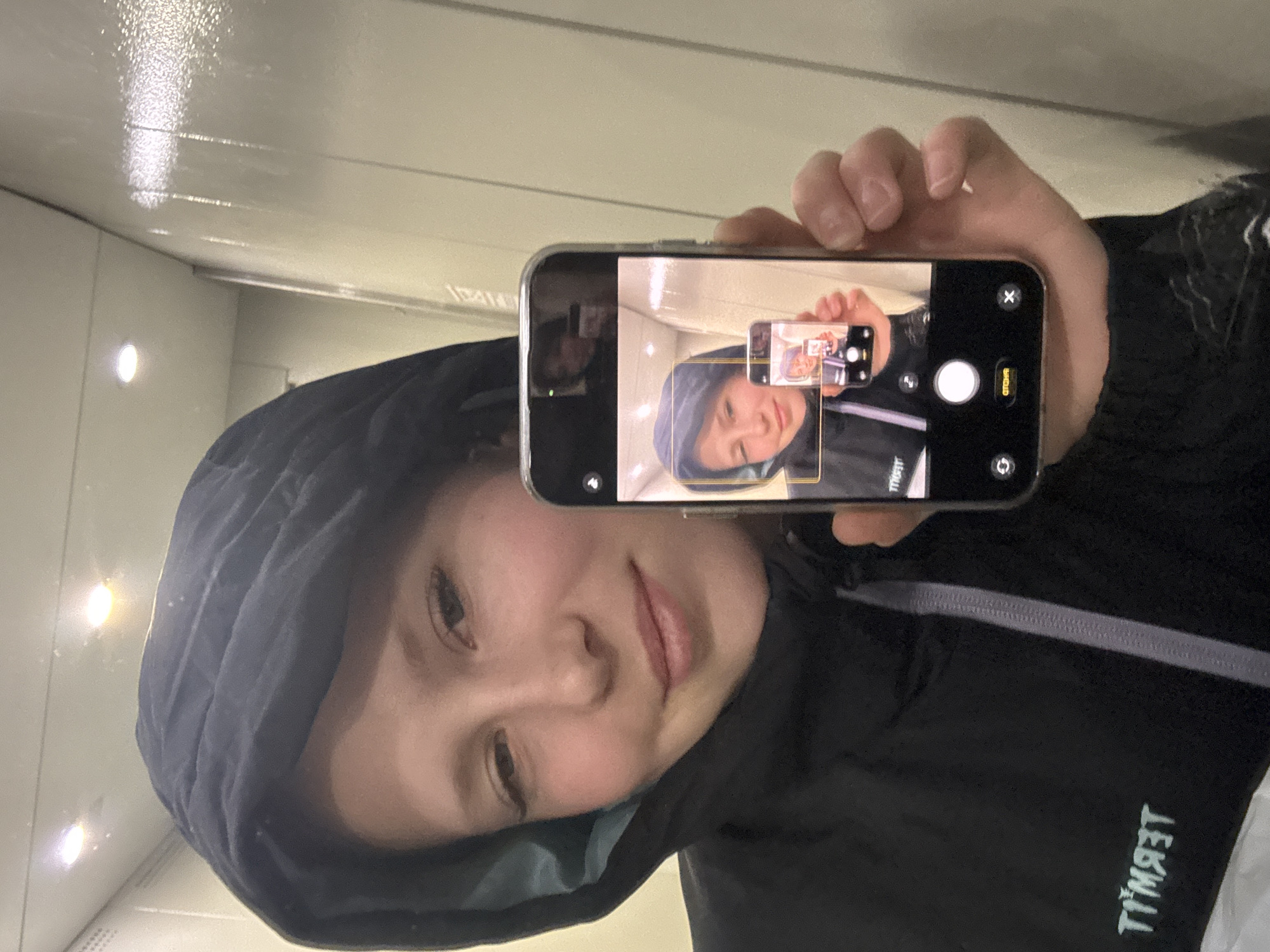 This media file presents a rotated close-up selfie of a young individual captured in a reflective surface. The subject is seen from the chest up, wearing a dark hooded jacket with the hood pulled over their head. Their expression is neutral to slightly content, and their gaze is directed towards the camera.
The central element is a silver-edged smartphone, held by the person's right hand. The phone's screen displays its camera application, showing a live preview of the person taking the selfie, creating a recursive "Droste effect" with an image within an image. Visible on the phone screen's interface are a large circular shutter button, a 'cancel' option, and controls for flash and camera switching.
The background consists of light-colored, possibly reflective, smooth surfaces that resemble walls or panels in an indoor setting. Straight lines and seams run horizontally across these surfaces. Several small, circular light sources are visible in the upper left, casting bright reflections and glares.
Visible text includes "ОТМЕНА" (OTMENA), meaning "CANCEL," on the phone's camera interface. On the person's jacket, a mirrored word, appearing as "TIMЯЯТ," is visible, likely a brand or logo. The image is oriented sideways, with the person's head tilted to the right. The specific time of day or detailed location cues are not evident beyond an indoor environment.