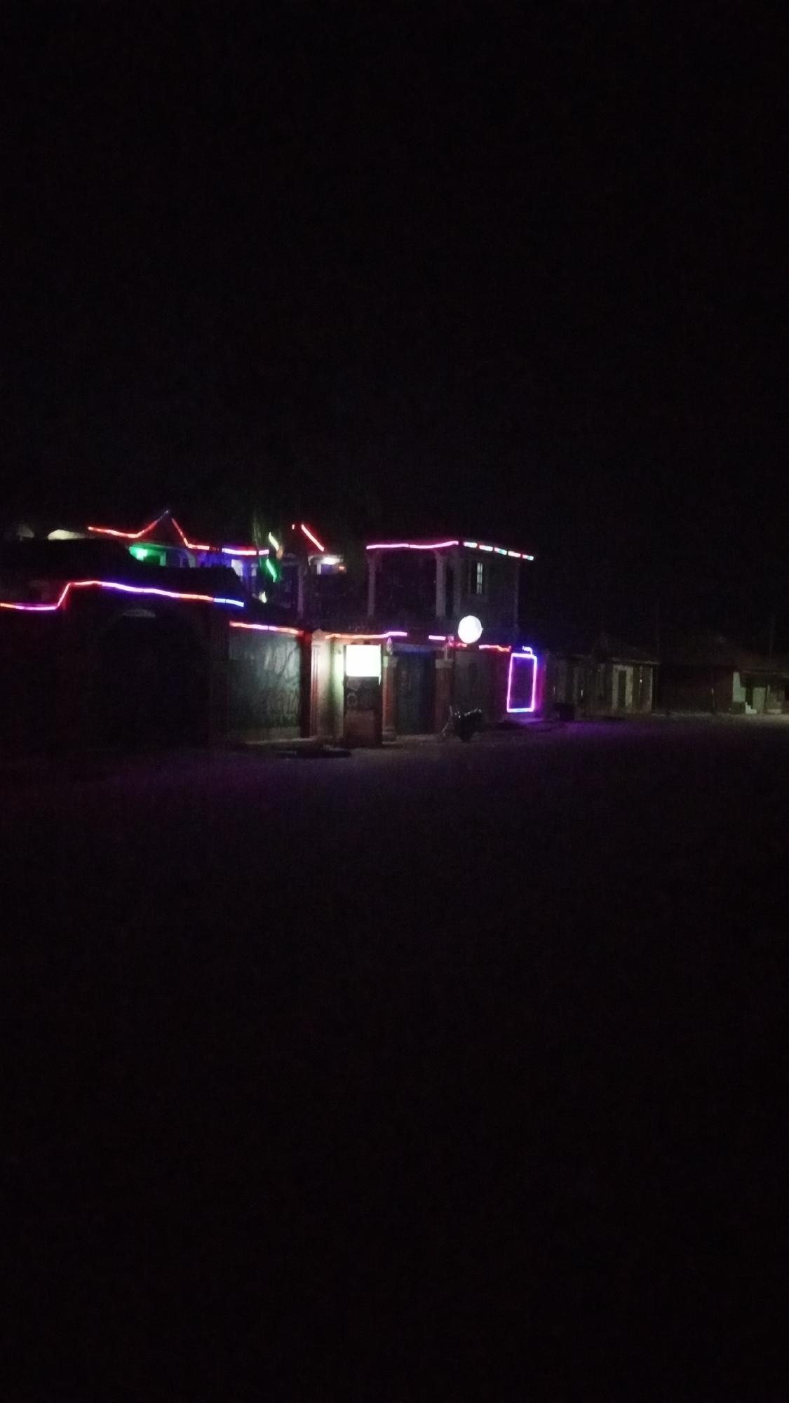 At night in Sabon Tasha, Nigeria, a building is decorated with neon lights. The lights outline the roof and parts of the walls in various colors, including red, green, blue, and purple. There is a round white sign on the front of the building, and a motorcycle is parked nearby. Other buildings are visible in the background, though less distinctly lit. The foreground is dark, suggesting a street or open area.