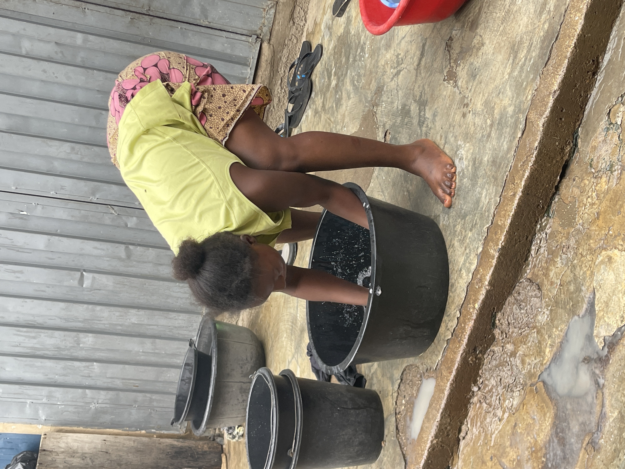 A young girl stands outdoors, leaning over a large black bucket, her arm submerged in water within the bucket, seemingly engaged in washing. She is wearing a yellow shirt and a skirt patterned with brown and pink. Her hair is tied up in a bun. Next to the large bucket are two smaller, black plastic buckets and a grey plastic bucket. A red bucket is visible in the top right corner of the image. The ground appears to be concrete with patches of discoloration. In the background is a corrugated metal wall, and a pair of black flip-flops. The photo was taken in Nyanya, Nigeria.