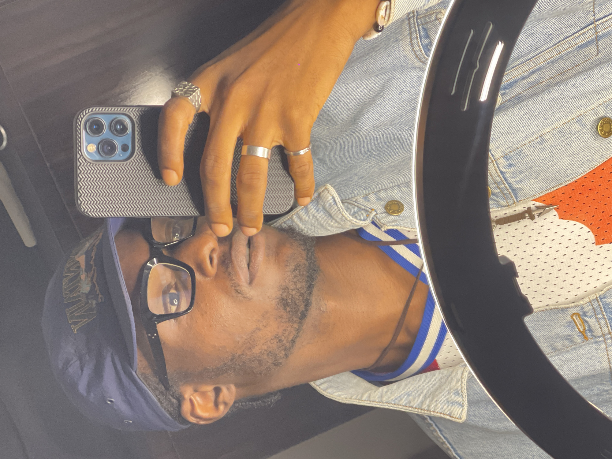 A person, appearing to be male with dark skin and facial hair, is reflected in a mirror. He is wearing a dark blue baseball cap with a gold emblem, black-rimmed glasses, a light blue denim jacket over a white mesh jersey with orange and blue accents, and several silver rings on his fingers. He is holding a smartphone in his right hand, taking a selfie. A ring light is visible in the foreground, partially obscuring the lower portion of the image. The background is dark and reflects the environment. The location is not specified in the image.