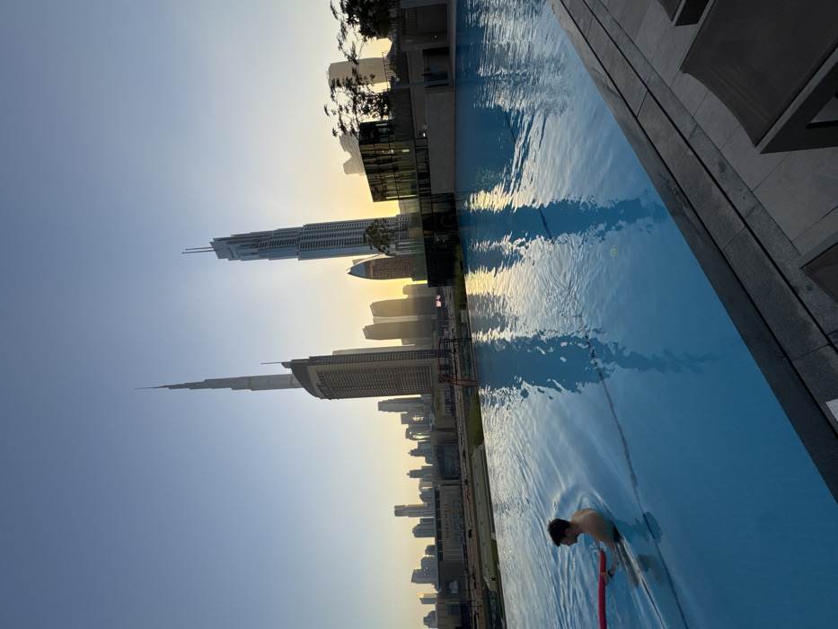 The image displays an outdoor infinity-edge swimming pool at dusk or dawn in Downtown Dubai, United Arab Emirates. The pool offers a direct view of the city's skyline, prominently featuring the Burj Khalifa centered in the background, accompanied by the Address Boulevard skyscraper to its right, and numerous other high-rise buildings. The "DUBAI MALL" signage is visible on a structure in the mid-ground, to the left of the Burj Khalifa.
In the foreground, a single adult male, light-skinned with dark hair, is partially submerged in the clear blue pool water, visible from the waist up. He is facing away from the camera, looking towards the cityscape, and holds a red foam noodle in his left hand. Ripples are visible in the water around him. The pool water reflects the golden-yellow light from the low sun, positioned behind the distant buildings, and the silhouettes of the skyscrapers.
The immediate right edge of the pool features dark tiled paving, with portions of two dark-fabric lounge chairs visible. Dark foliage from trees or bushes is present along the far right edge of the pool, contributing to the reflections in the water.