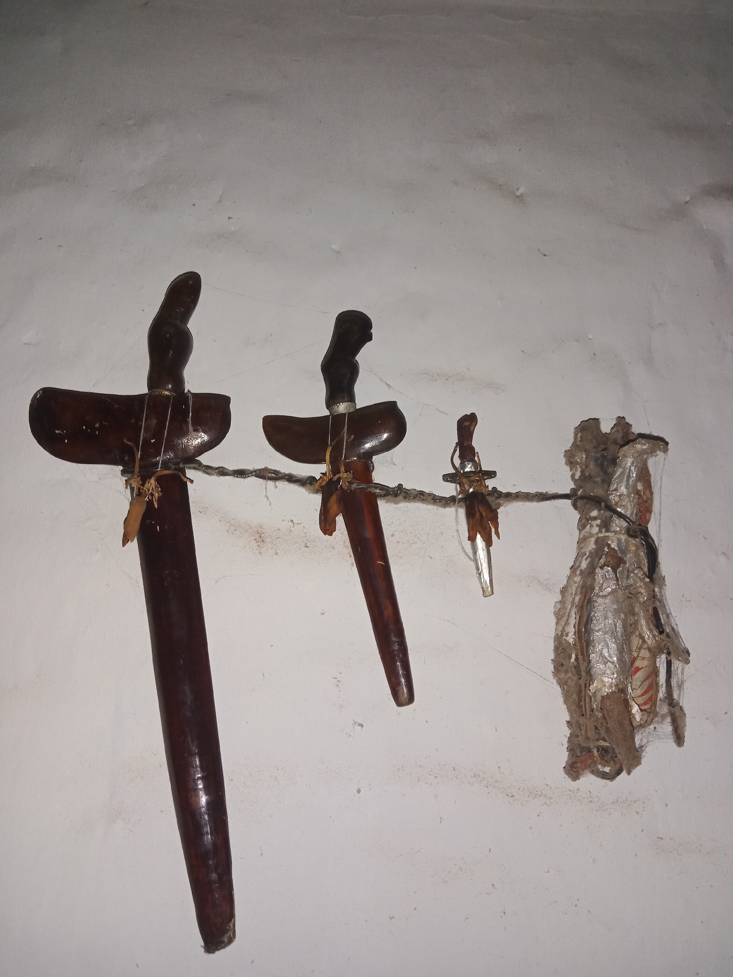 A wall in Pohsangit Leres, Indonesia, features three items suspended on a wire: two daggers and a bundle of what appears to be dried organic material. The daggers are positioned to the left, one larger than the other, both with dark wooden handles and attached to the wire by string. The larger dagger has a curved, dark brown handle and a prominent blade. The smaller dagger is similar in construction but shorter. To the right of the daggers, the wire holds a loosely tied bundle of what appears to be dried leaves, fibers, and possibly bone fragments, wrapped in some form of coarse fabric or netting. The wall itself is rough and textured, with visible signs of age and discoloration.