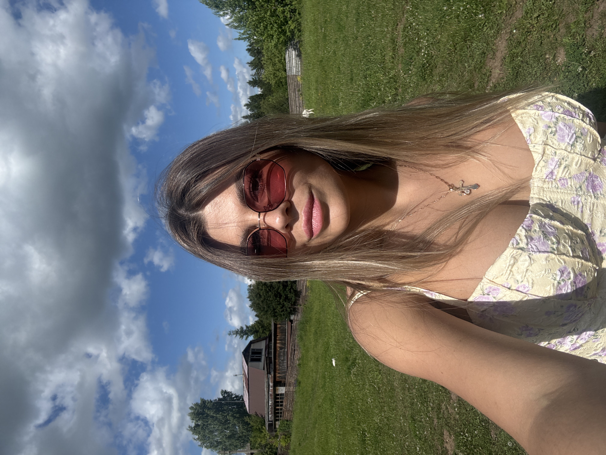 A young woman with blonde hair and pink-tinted sunglasses takes a selfie in a grassy field. She is wearing a yellow, floral-patterned top and a necklace with a cross pendant. Behind her, a wooden fence encloses a field with a rustic wooden house and trees. The sky is blue with scattered white clouds. The location is identified as Pastukhovo, Russia. A white animal, possibly a dog, is visible in the distance.
