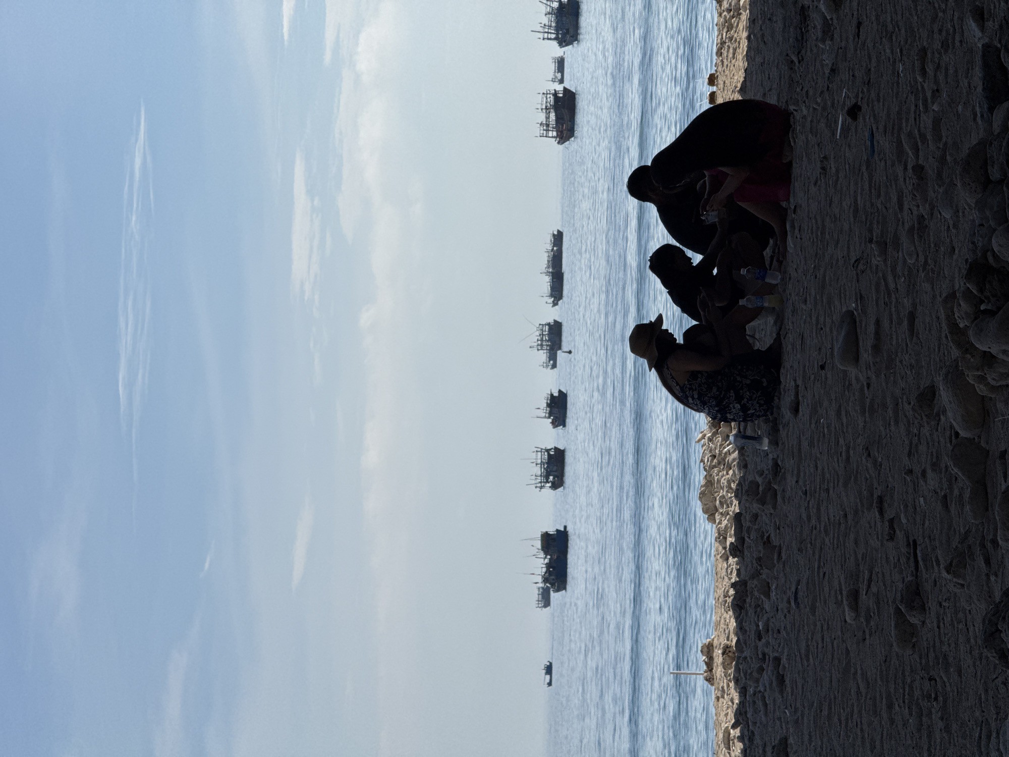 The image depicts a serene beach scene in Lý Sơn Island, Vietnam. In the foreground, a family of four is silhouetted against the ocean, engaged in an activity, possibly playing a game or interacting with their phones. The family is seated on the sandy beach, which is scattered with small stones and shells. Behind them, the calm blue ocean stretches towards the horizon, dotted with several fishing boats. The sky above is a pale blue, with a few wispy clouds, suggesting it is either early morning or late afternoon. The overall atmosphere is peaceful and tranquil, capturing a moment of leisure and connection amidst the natural beauty of the Vietnamese coast.