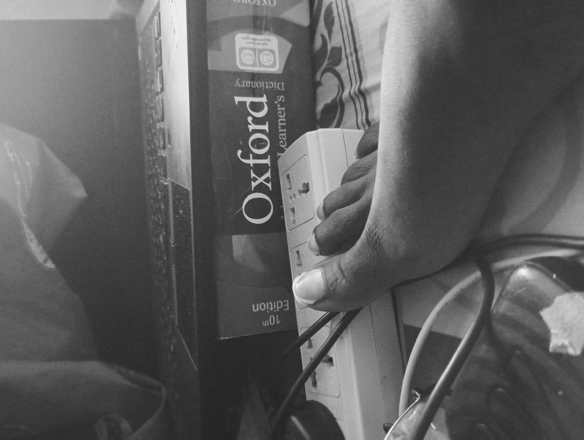 A black and white, low-angle, close-up shot captures a person's right foot resting on a white electrical power strip. The foot, with its toenails visible, is positioned on top of the power strip, covering some of the outlets. The power strip is connected to dark cables that snake around it. Behind the power strip, an open book with "Oxford Learner's Dictionary" printed on its cover is visible. To the left of the book, a laptop with its keyboard exposed is present. The background is obscured by dark, blurry shapes, possibly fabric or other objects. The overall scene appears to be indoors, with an emphasis on the intersection of technology and study materials, underscored by the presence of electrical components. The lighting is dim, creating a chiaroscuro effect, with harsh shadows and highlights. The image conveys a sense of casual, perhaps late-night, study or work.