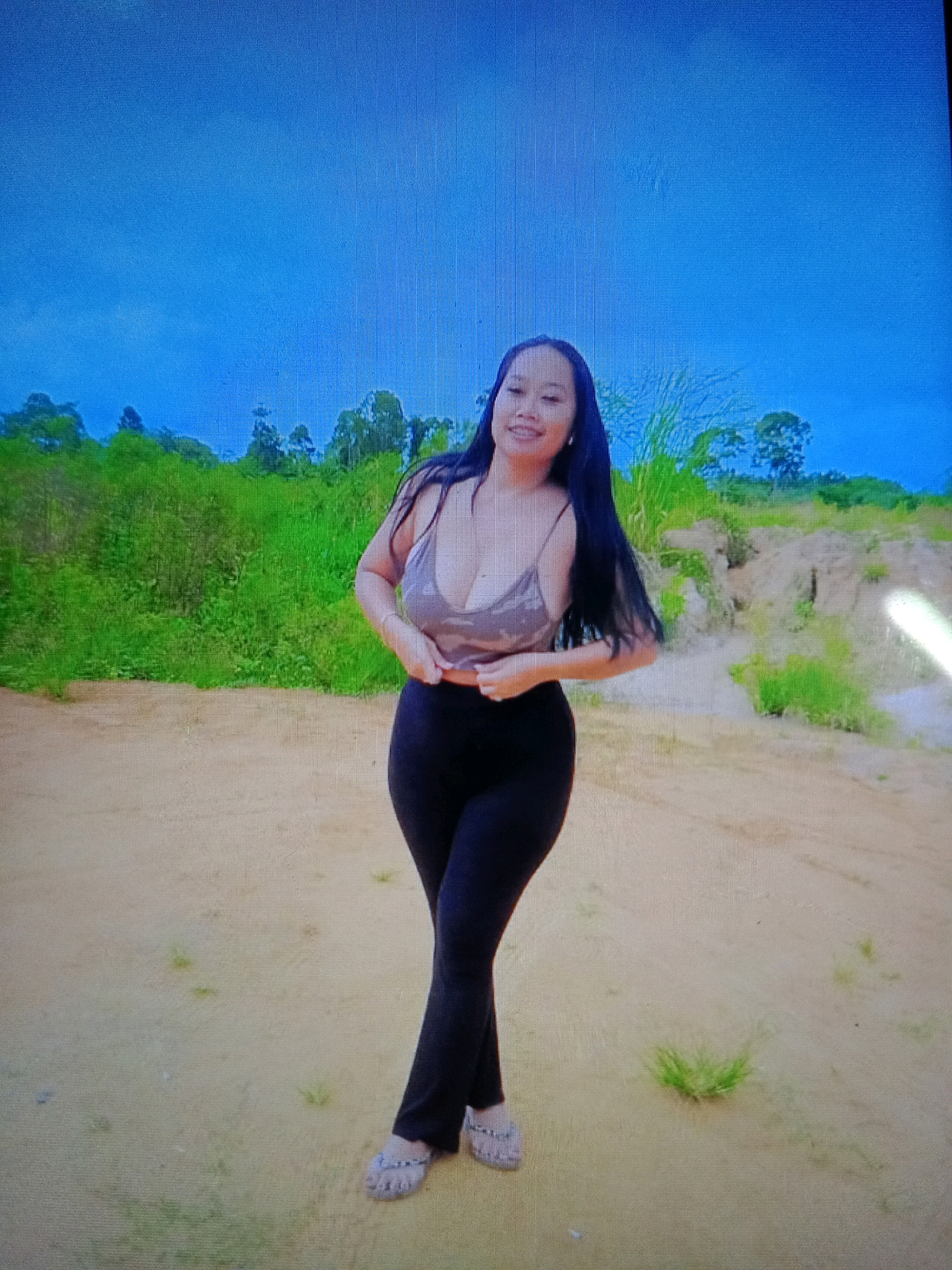 A young woman with long black hair stands in a sandy, outdoor location with green vegetation and a blue sky in the background. She is wearing a light-colored, spaghetti-strap top and dark, form-fitting pants. Her hands are on her stomach, and she is smiling at the camera. The ground appears to be a mix of sand and sparse green grass. The overall impression is of a casual outdoor portrait. The location is described as being in the city of Noord, Suriname.