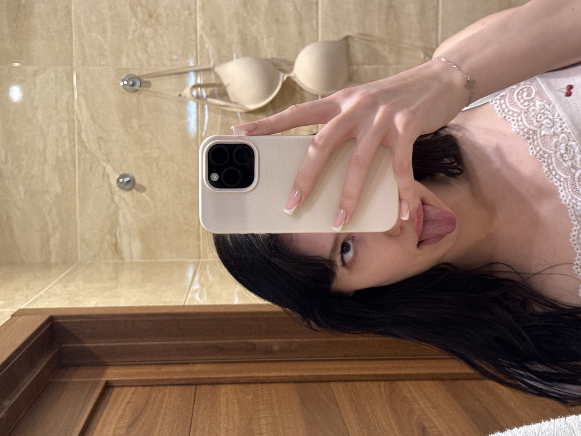 The image shows a young woman taking a selfie in what appears to be a bathroom. She has long, dark hair and is sticking her tongue out. She is wearing a white camisole with lace trim. In the background, there is a wooden door on the left and tiled walls with two hooks on which a beige bra is hanging. The image is not taken in Khimki, Russia.