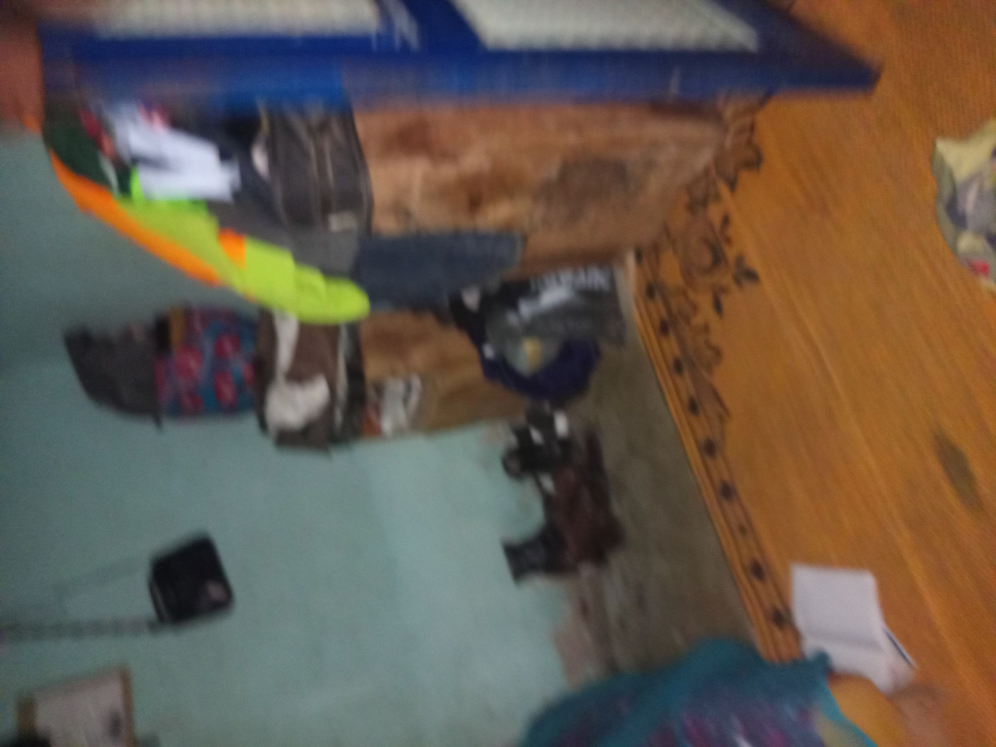 The image depicts an indoor scene with a light blue wall and a wooden floor with a patterned rug. In the background, there is a dark-colored bag hanging on the wall, and a stack of items including a colorful patterned box, a black bag, and some fabric. To the right, a tall, blue object, possibly a cabinet or shelf, is visible. In the middle ground, there appear to be some dark-colored objects on the floor, possibly animal figures or toys. In the foreground, the wooden floor and a portion of the rug are prominent. The image is blurry, making it difficult to discern specific details. The overall impression is that of a cluttered room. The location is identified as Jalingo, Nigeria, though there are no specific visual cues in the image to confirm this location.