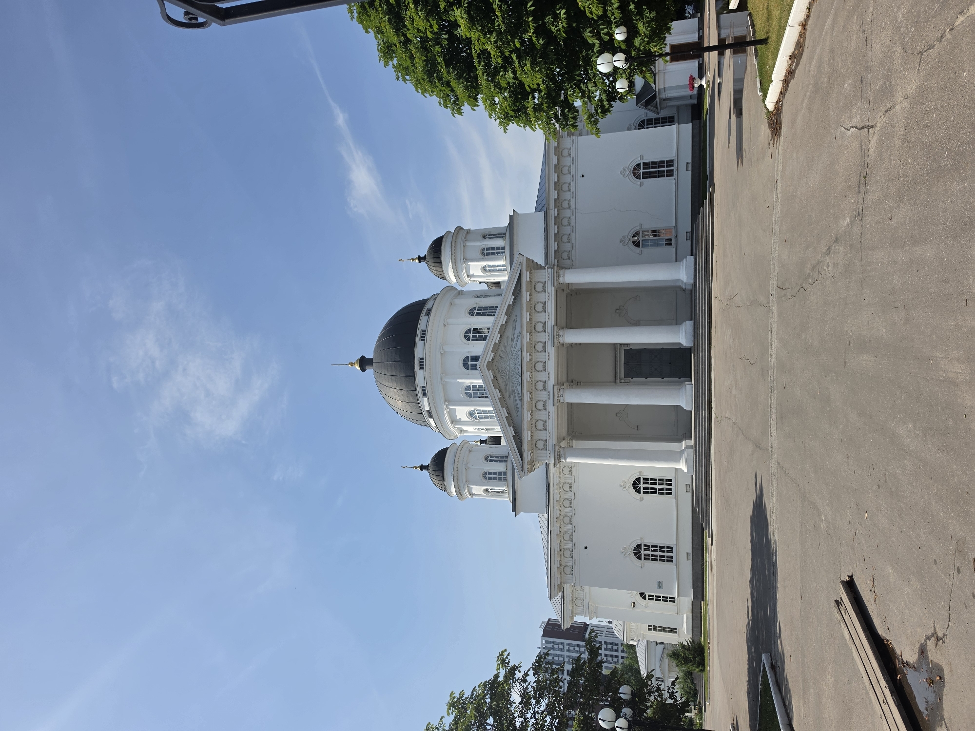 The image shows a large, white neoclassical church with a central black dome and two smaller black domes on either side. The church has a grand entrance with a portico supported by several tall columns. The facade features arched windows and ornate architectural details. The church is set against a clear blue sky with some wispy clouds. In the foreground, there is a paved area with some cracks and debris. Trees line the sides of the church, and a modern building can be seen in the background to the left. The overall scene depicts a well-maintained public space in a city. This building is the Alexander Nevsky Cathedral located in Nizhny Novgorod, Russia.