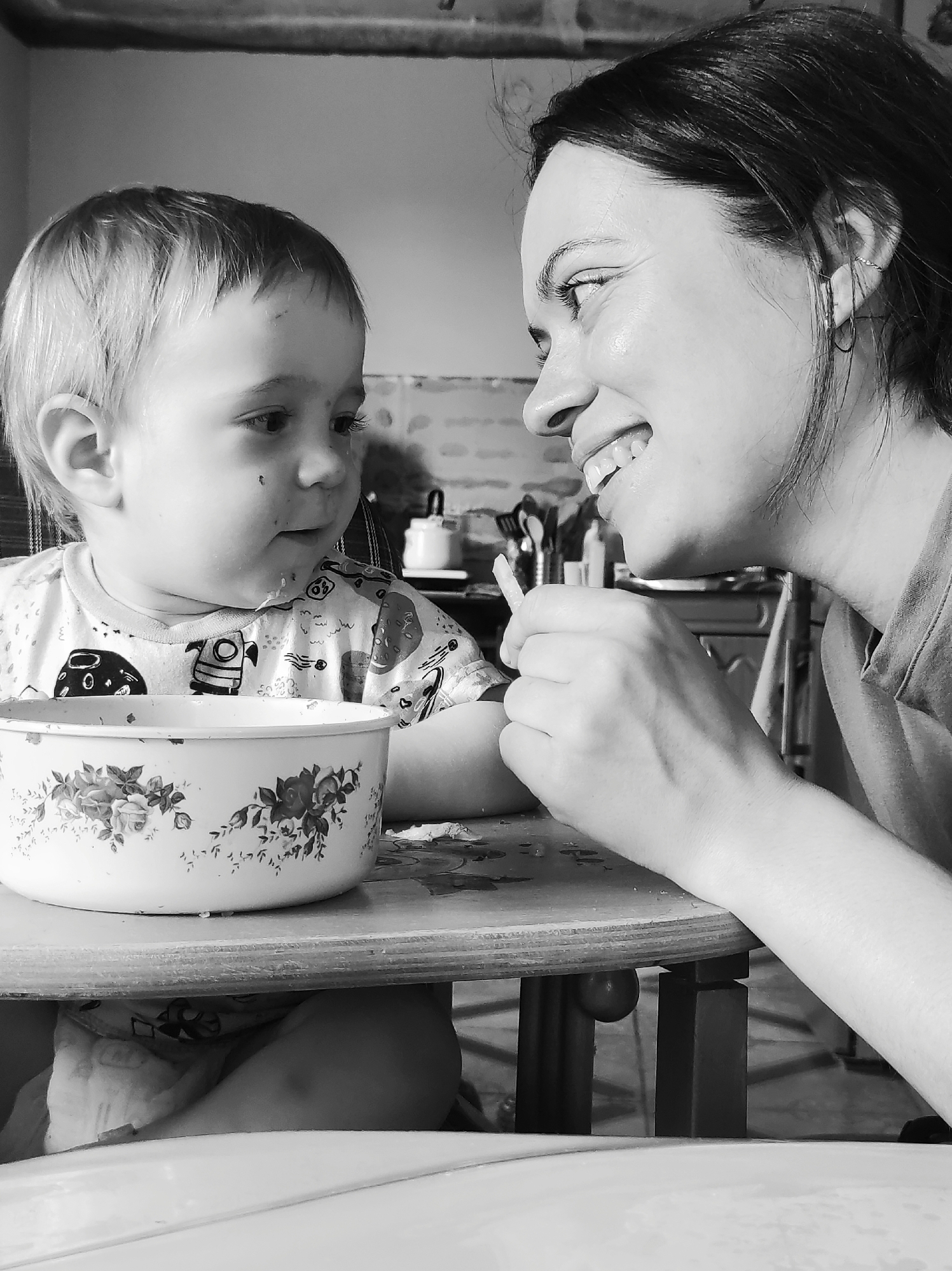 The image, a black and white photograph, depicts a woman and a child at a table. The woman is on the right side of the frame, her face visible in profile. She is smiling and looking at the child. The child, a young boy, is on the left side of the frame, seated at the table with a large bowl in front of him. The bowl is decorated with floral patterns. The child is looking at the woman. The woman is holding something in her hand, possibly food, offering it to the child. The table is wooden, and there are objects visible in the background, including kitchen utensils. The image appears to be taken indoors, possibly in a kitchen. There is no information that indicates the location is in the city of Фоки, Russia.