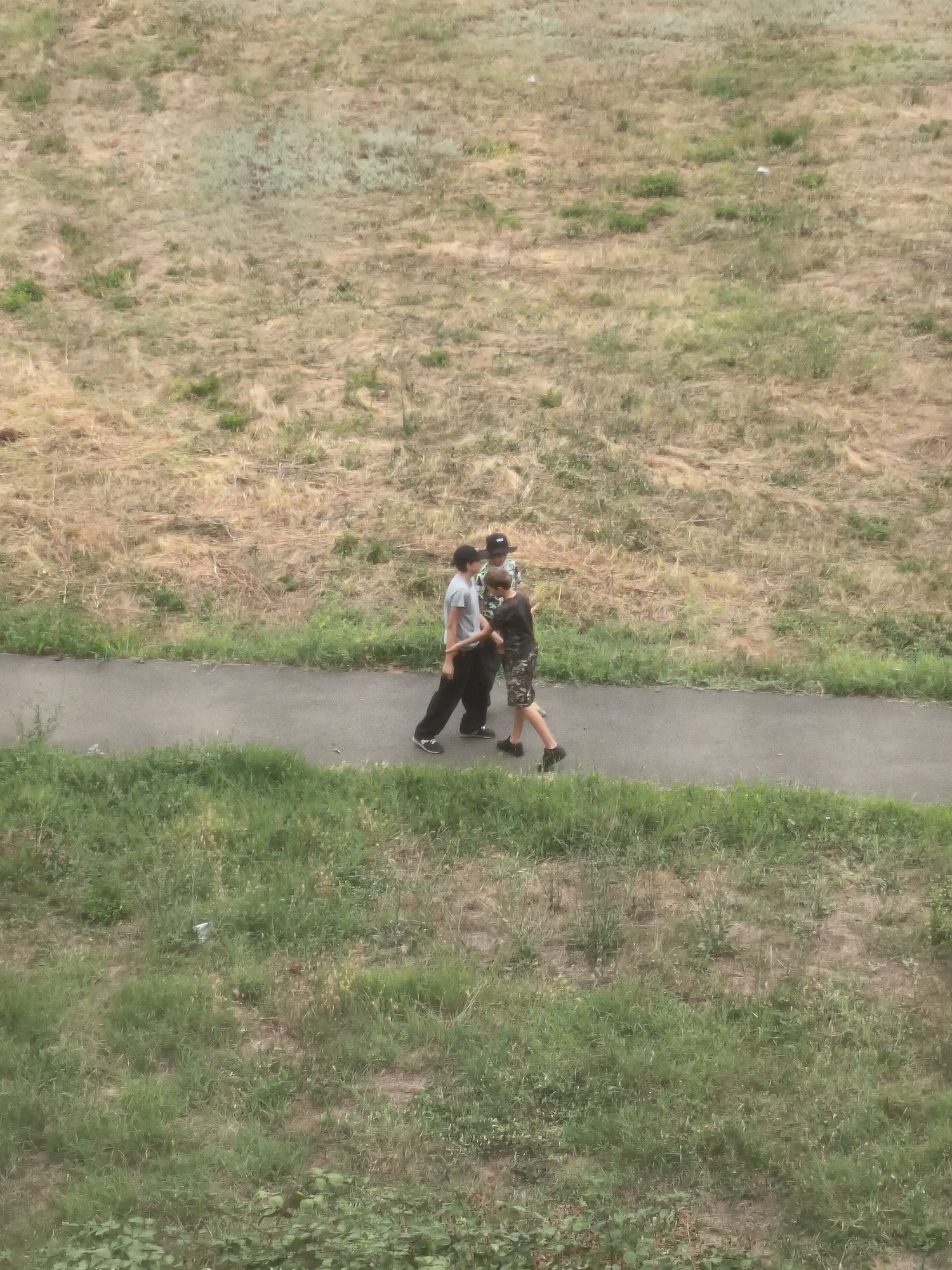 In Izmail, Ukraine, two young males are walking on a paved path that runs through a grassy area. The male on the left is wearing a grey t-shirt and black pants, and the male on the right is wearing a camouflage t-shirt and camouflage shorts. They are walking side-by-side, facing away from the camera. The path is bordered by dry grass and sparse green vegetation on one side, and by green grass on the other. The background is a dry, textured hillside covered in sparse vegetation. The lighting suggests it is daytime, possibly late afternoon or early morning, given the shadows.
