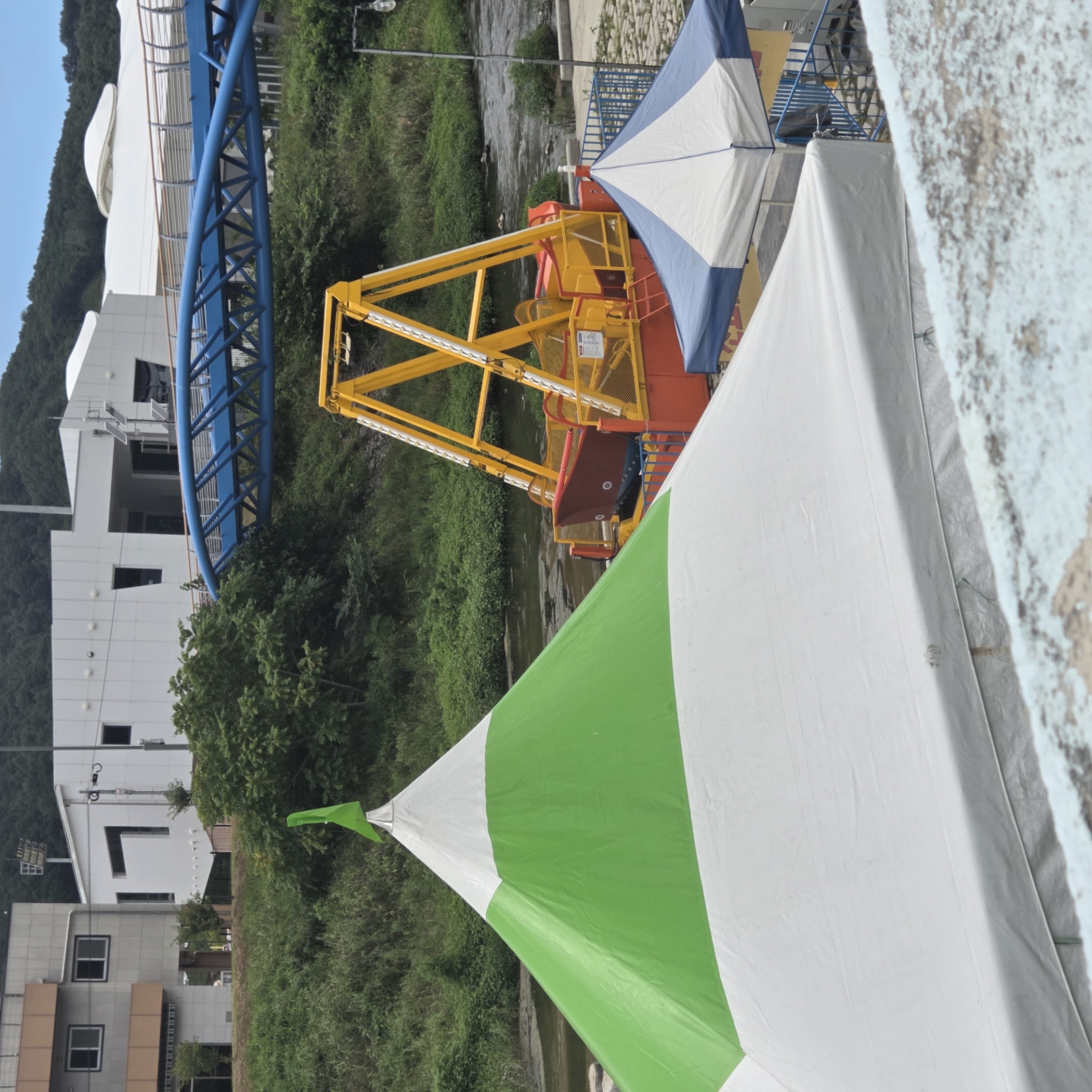 The image shows an outdoor scene near a river in Dunnae-myeon, South Korea. In the foreground, a large white tent with a green conical top and a green triangular flag is visible. To the right, a blue and white striped tent is partially visible. In the middle ground, a bright yellow amusement park ride, possibly a swing ride or a pirate ship, is situated near the riverbank. The ride has a red gondola and orange base. Behind the ride, a blue pedestrian bridge arches over the river. The background features green vegetation and hills, with a few buildings, including a light-colored structure with windows and a dark-colored building, visible on the left side of the frame. The sky is overcast with blue patches. The scene appears to be set up for an outdoor event or festival.