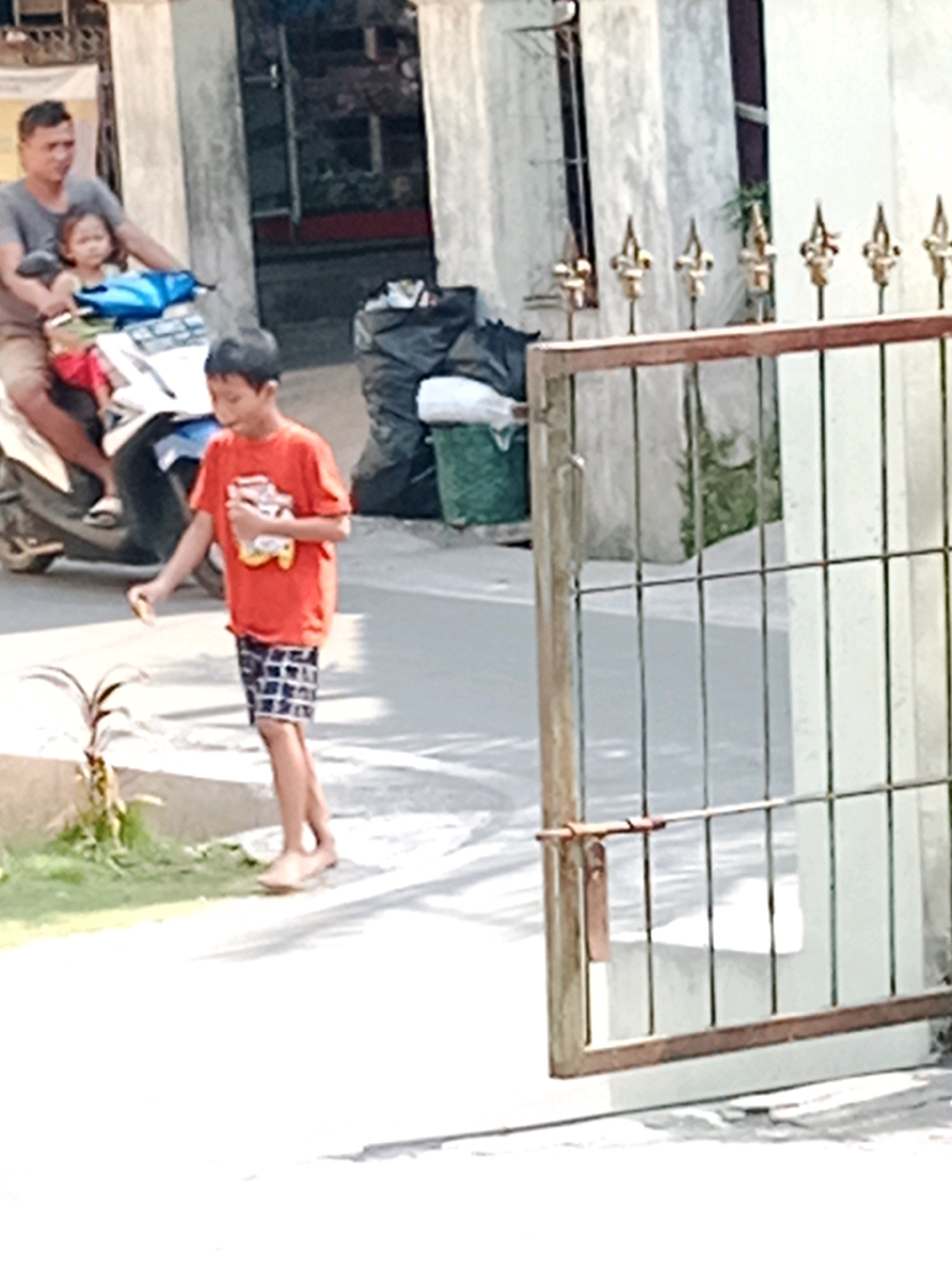A man, a child, and a young boy are in the foreground and middle ground of this outdoor scene, which is likely located in Waru, Indonesia. The man, wearing a gray t-shirt and shorts, is seated on a white scooter, with the child, a young girl wearing a pink shirt and red pants, on his lap. The boy, in an orange t-shirt and plaid shorts, is standing to the right of the scooter, holding a small object in his right hand. He appears to be interacting with the environment. Behind the boy are several large black garbage bags. In the background, there is a white wall and a metal fence with decorative elements. The ground is paved, and there are some plants visible to the left of the boy. The overall lighting suggests it is daytime.