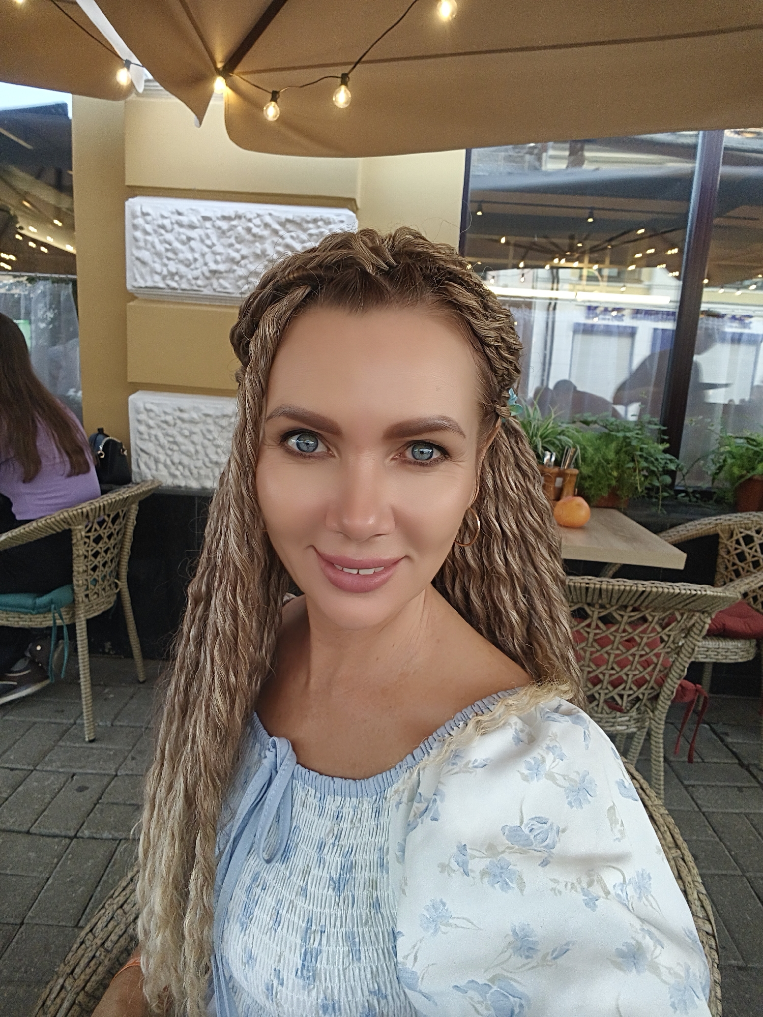 A woman with blonde hair in braids and extensions, blue eyes, and a light blue and white floral off-the-shoulder top smiles at the camera. She is sitting at a wicker chair at an outdoor cafe. In the background, a woman with long brown hair is seated at another table, and plants are visible on a table. The overall setting appears to be a sunny day. This image may have been taken in Yaroslavl, Russia.