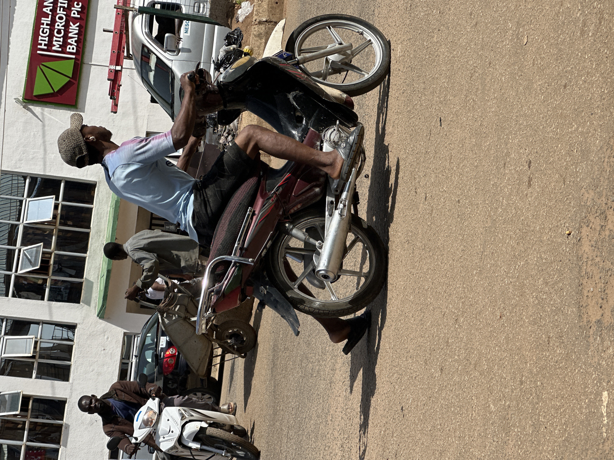 The image depicts a street scene in Bukuru, Nigeria. In the foreground, a man wearing a light blue t-shirt, dark shorts, a baseball cap, and sandals is riding a red and black motorcycle. He has earbuds in his ears. Behind him, a white motorcycle is parked, and a man in a brown jacket is seated on it, looking forward. Further in the background, a dark-colored car is parked. To the right of the man on the motorcycle, a silver van is visible with the "NESO" logo on its side. Behind the van, a building with multiple windows can be seen, and a red sign with green triangular shapes and the text "HIGHLAND MICROFINANCE BANK Plc" is mounted on the building's exterior. The road surface is asphalt with visible tire marks and shadows.