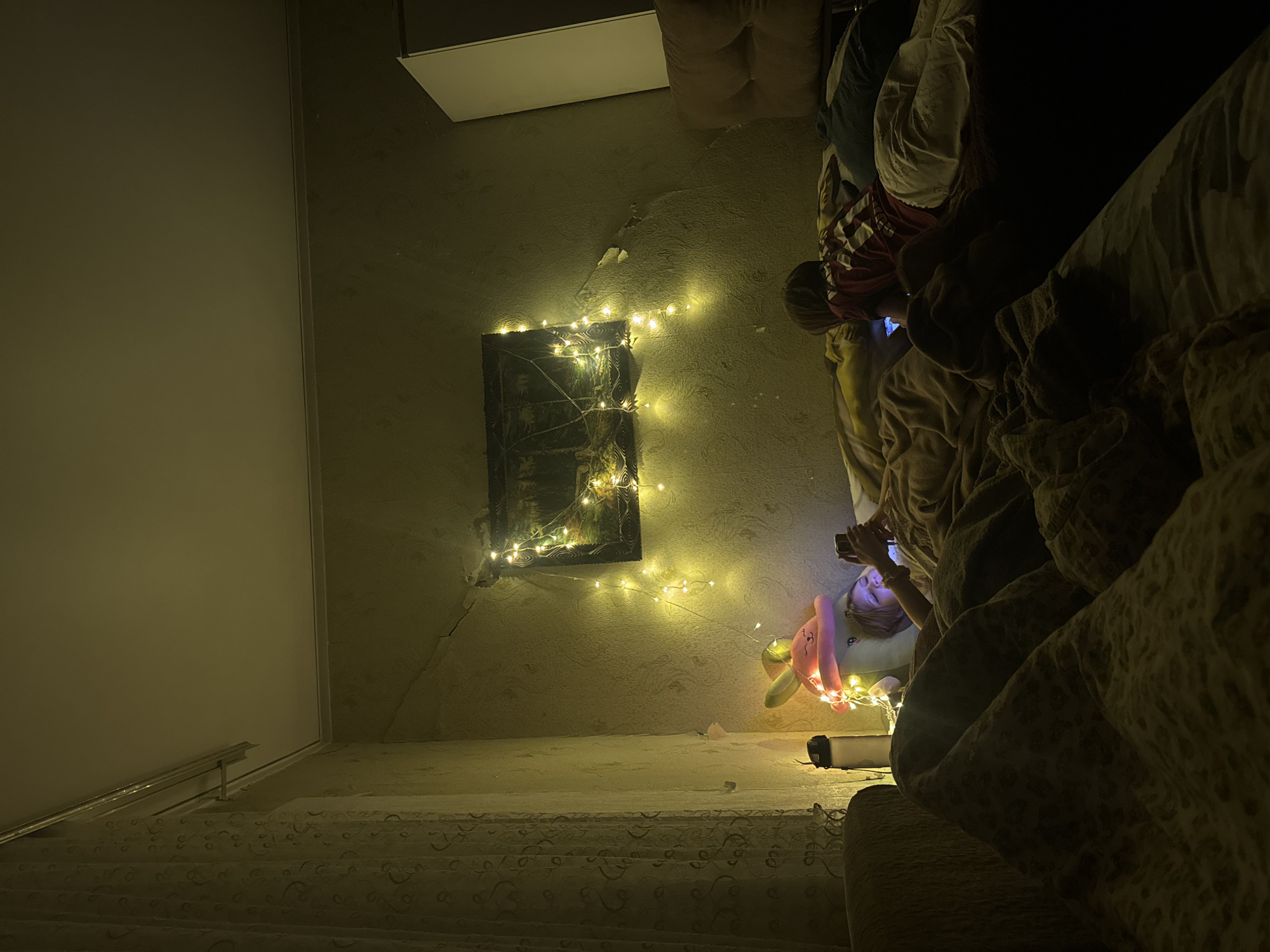 The image depicts an indoor scene, likely a bedroom, with two individuals lying on a bed. The room is dimly lit, with the primary light source appearing to be string lights draped around a framed picture on the wall. The picture itself appears to be a landscape or nature scene.
To the left, a person is lying on their side, looking at a mobile phone. They are partially covered by blankets and a large, plush toy with bunny ears, also illuminated by the string lights. Next to them, a water bottle stands upright.
To the right, another person, a child, is also lying on the bed, facing away from the camera and looking at a mobile phone. They are wearing a red and white striped shirt and light-colored pants.
The room's walls have a textured wallpaper, and there's evidence of cracking or damage in some areas. A curtain is visible on the left side of the frame. The overall impression is one of a cozy, dimly lit evening at home. The location is identified as Shakhty, Russia.
