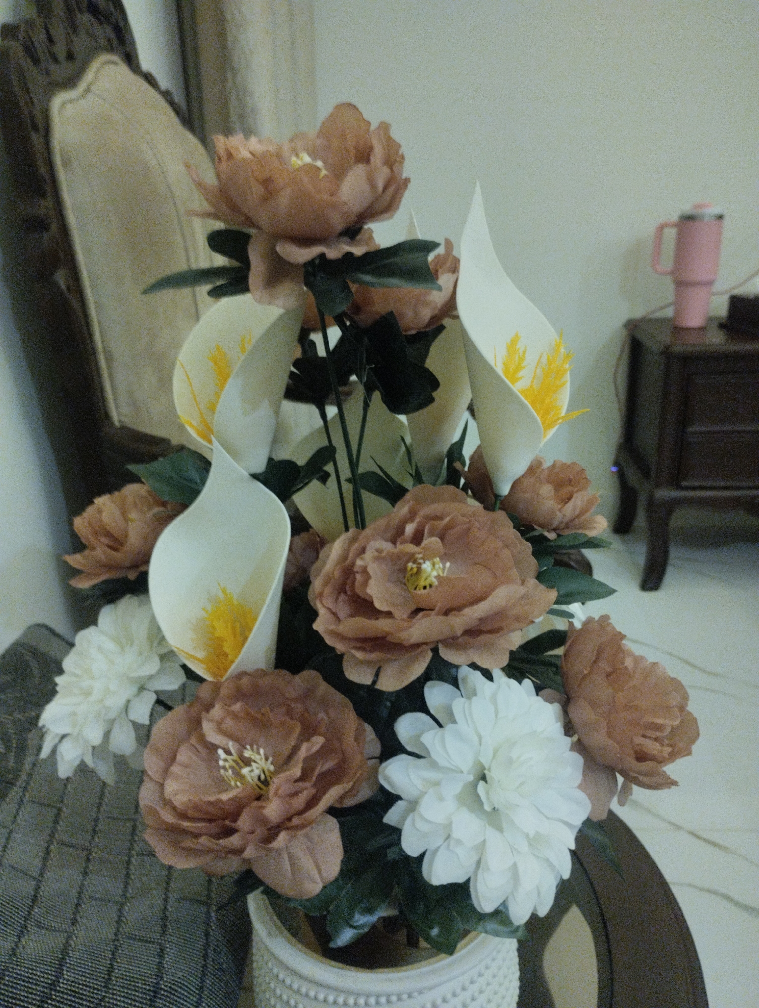 In an indoor setting, a floral arrangement is prominently displayed. The bouquet features a mix of large, taupe-colored peonies and white dahlias. Interspersed among the flowers are elegant white calla lilies with yellow stamen. The arrangement is housed in a textured white vase. To the left, a portion of a light-colored upholstered chair with carved dark wood is visible. In the background, to the right, a dark wooden side table holds a pink tumbler and other indistinct items. The scene appears to be situated in a living space.