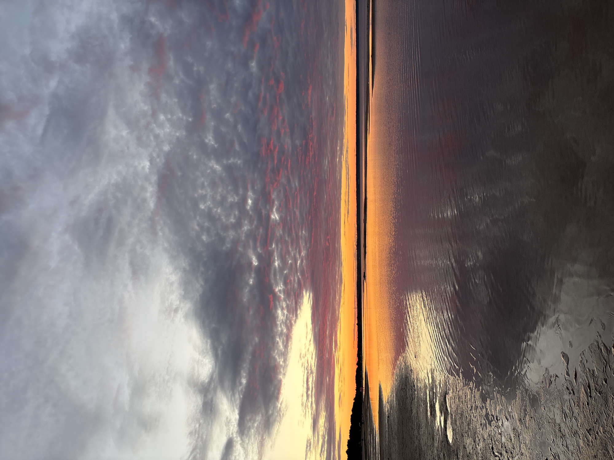 The image captures a serene sunset scene at a beach in Kokkorevo, Russia. The sky is adorned with dramatic clouds, a mix of grey and pink hues, illuminated by the warm glow of the setting sun. The horizon is marked by a strip of orange and yellow light, transitioning into the darker tones of the sky. Below, a calm body of water reflects the vibrant colors of the sunset, creating a mirror-like effect. The foreground features a sandy beach with shallow water receding from the shore, revealing wet sand with small puddles. On the left side of the beach, a dark silhouette of trees and a few indistinct figures suggest a distant treeline and possible human presence. The overall atmosphere is peaceful and visually striking, with the interplay of light and shadow on the water and sand adding depth to the composition.